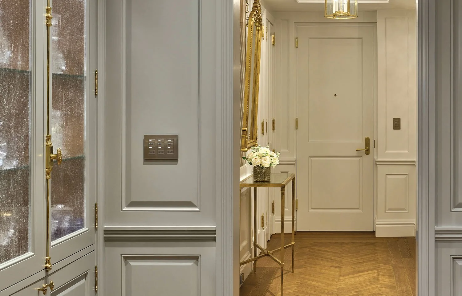 Elegant foyer with paneled cream walls and brass accents, featuring a glass front cabinet with textured glass, a Lutron Keypad on the wall, a gold framed concole table holding white roses, a pendant light above, and a herringbone wood floor leading t