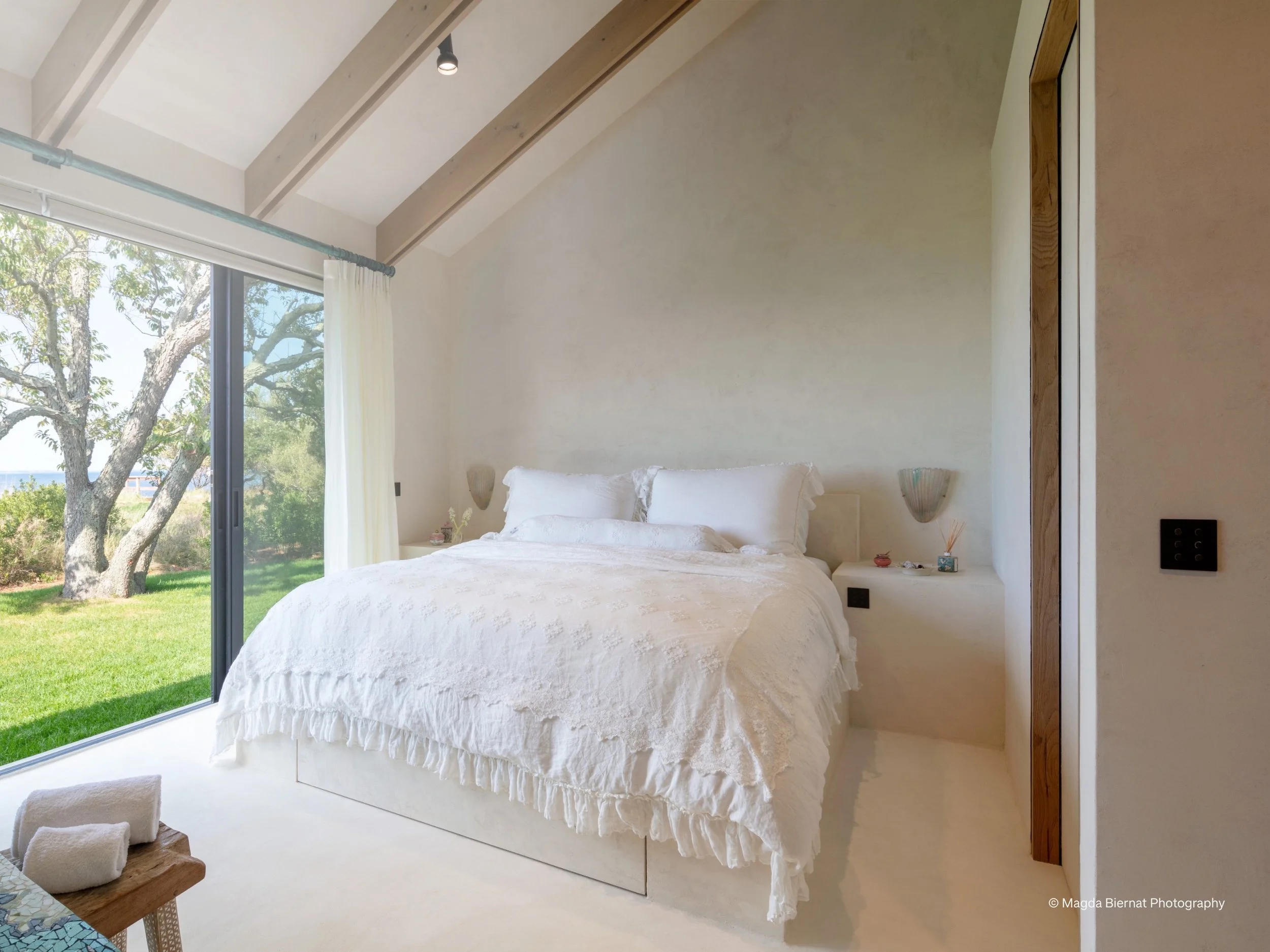 A tranquil modern bedroom with Lutron motorized sheer shades that filter natural light while framing outdoor views. The space features a vaulted ceiling with exposed beams, soft neutral walls, and layered white bedding, creating a calm, spa-like retr
