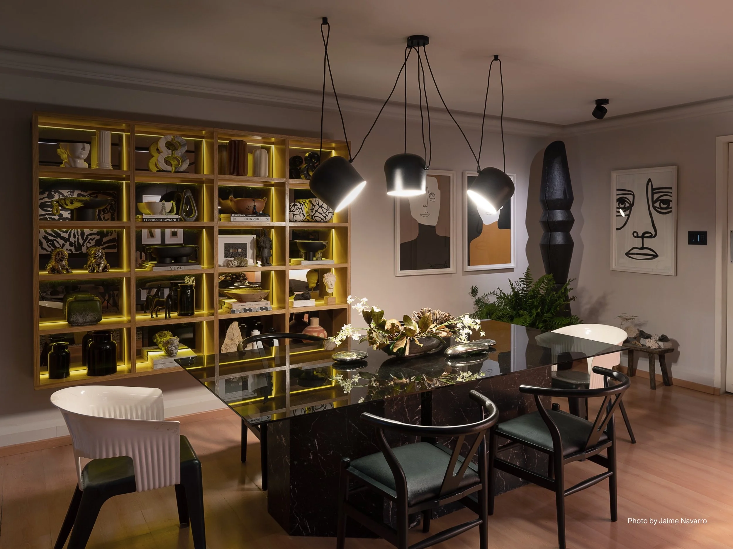 A bold, modern dining room illuminated by Lutron smart lighting, creating a striking balance of drama and refinement. The space features a sleek black marble dining table surrounded by mixed modern chairs. Above, a cluster of contemporary pendant lig