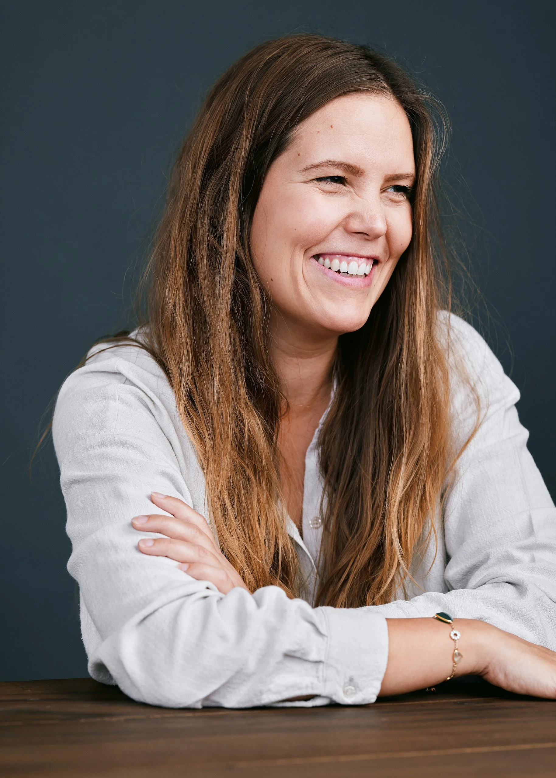 En smilende kvinde med langt, brunt, bølget hår, iført en beige hørskjorte, sidder ved et bord mod en mørk blå baggrund.