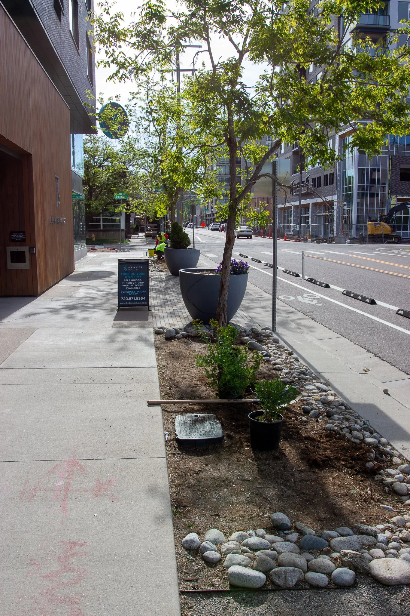 Sidewalk with newly planted shrubs and trees, construction work ongoing on the street, and an electronic sign advertising a tour near the building.