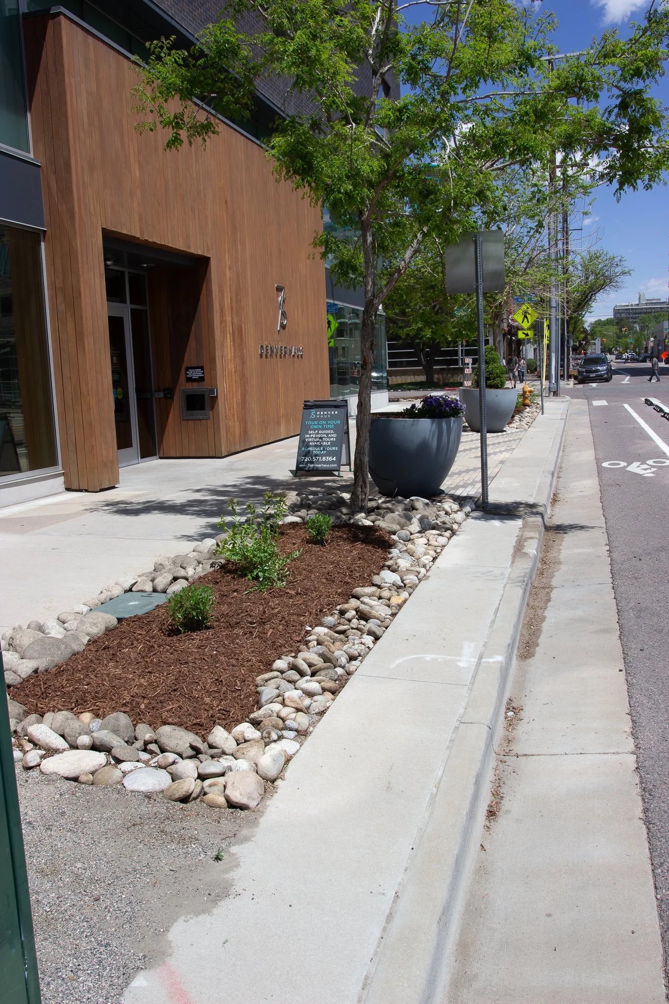 Sidewalk scene with a modern building, plants, and trees along the street, with cars and pedestrians in the background.