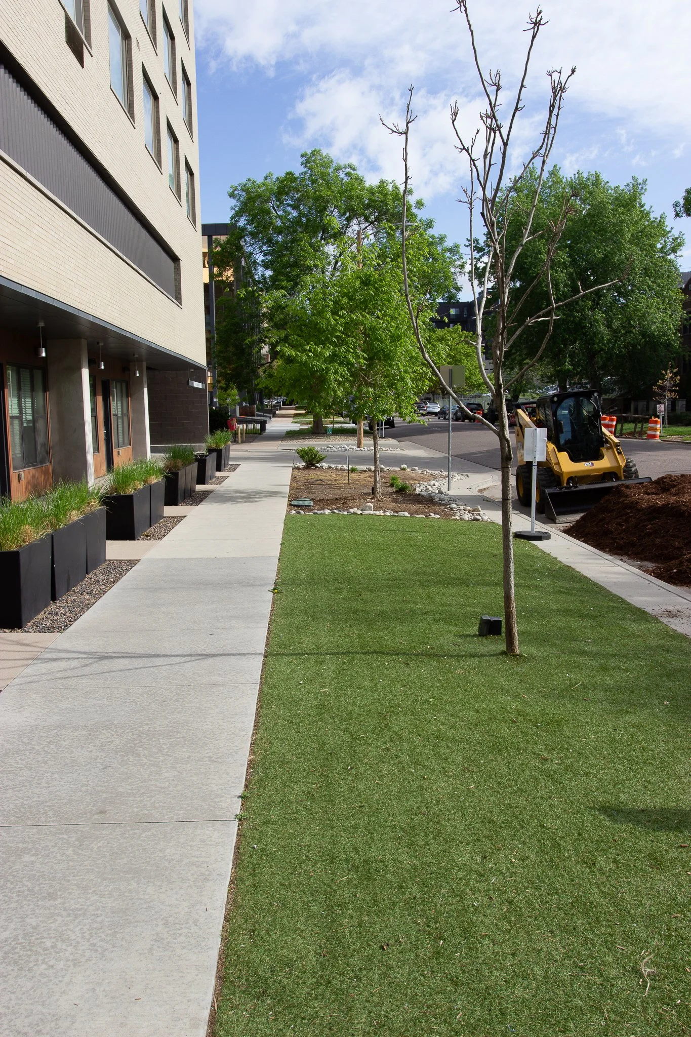 Sidewalk lined with planters and young trees outside a modern apartment building, with some trees being planted and a small yellow construction vehicle nearby under a partly cloudy sky.