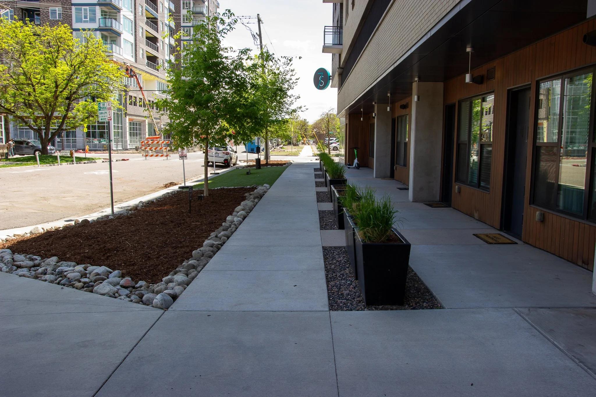 Sidewalk with small trees and planters along a modern multi-story building in an urban area.