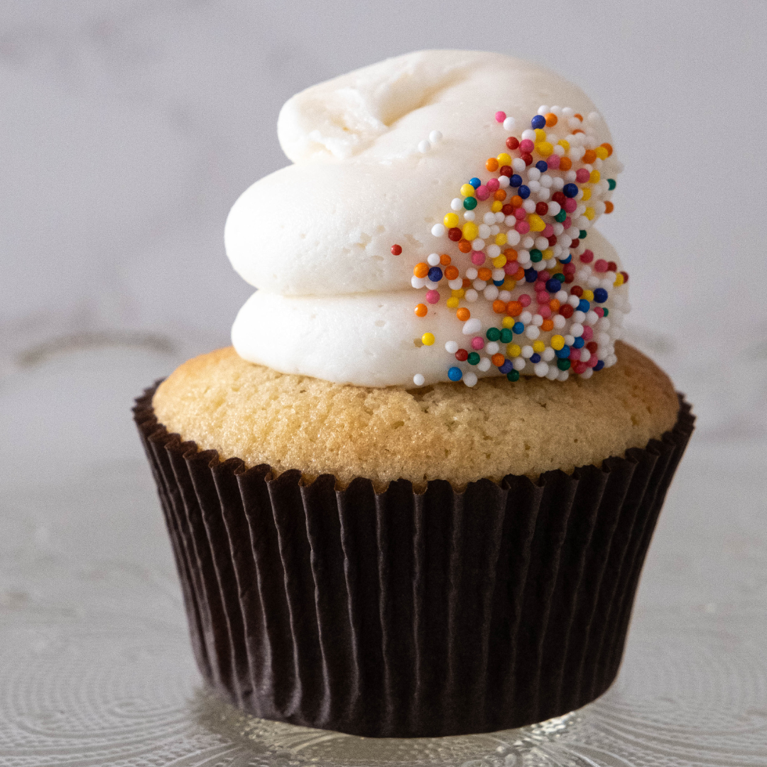 Close-up of a vanilla cupcake with a dark brown wrapper, topped with white frosting and colorful round sprinkles.