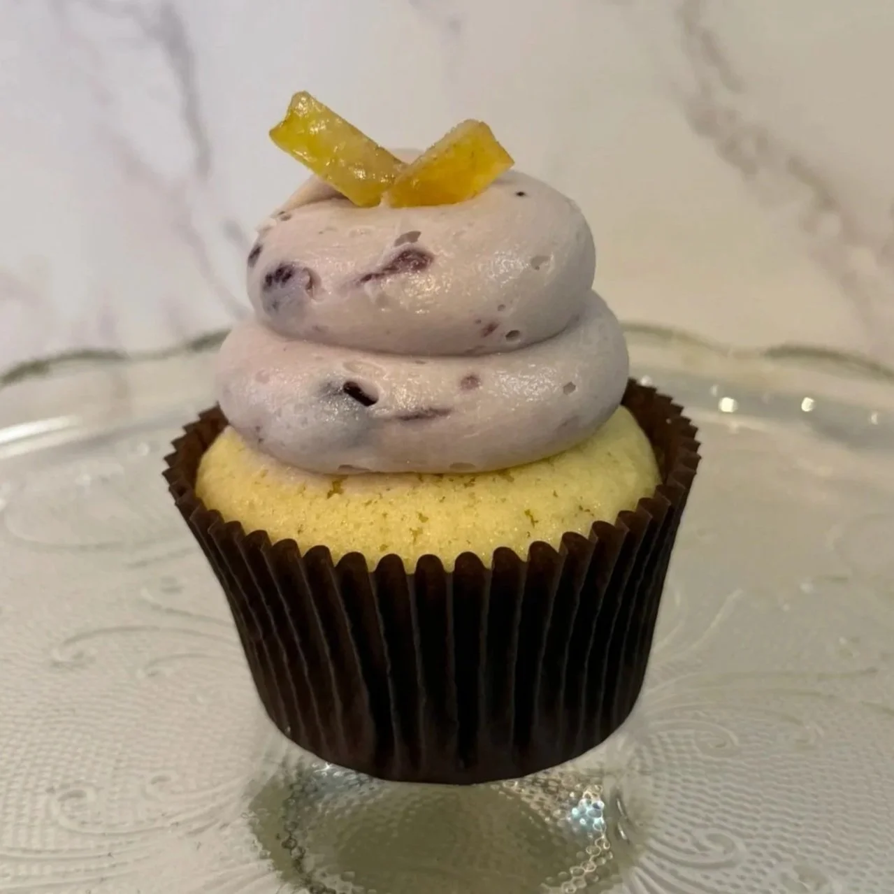 Close-up of a cupcake with purple frosting and lemon peel on top piece of orange candy on top, in a black paper cup.