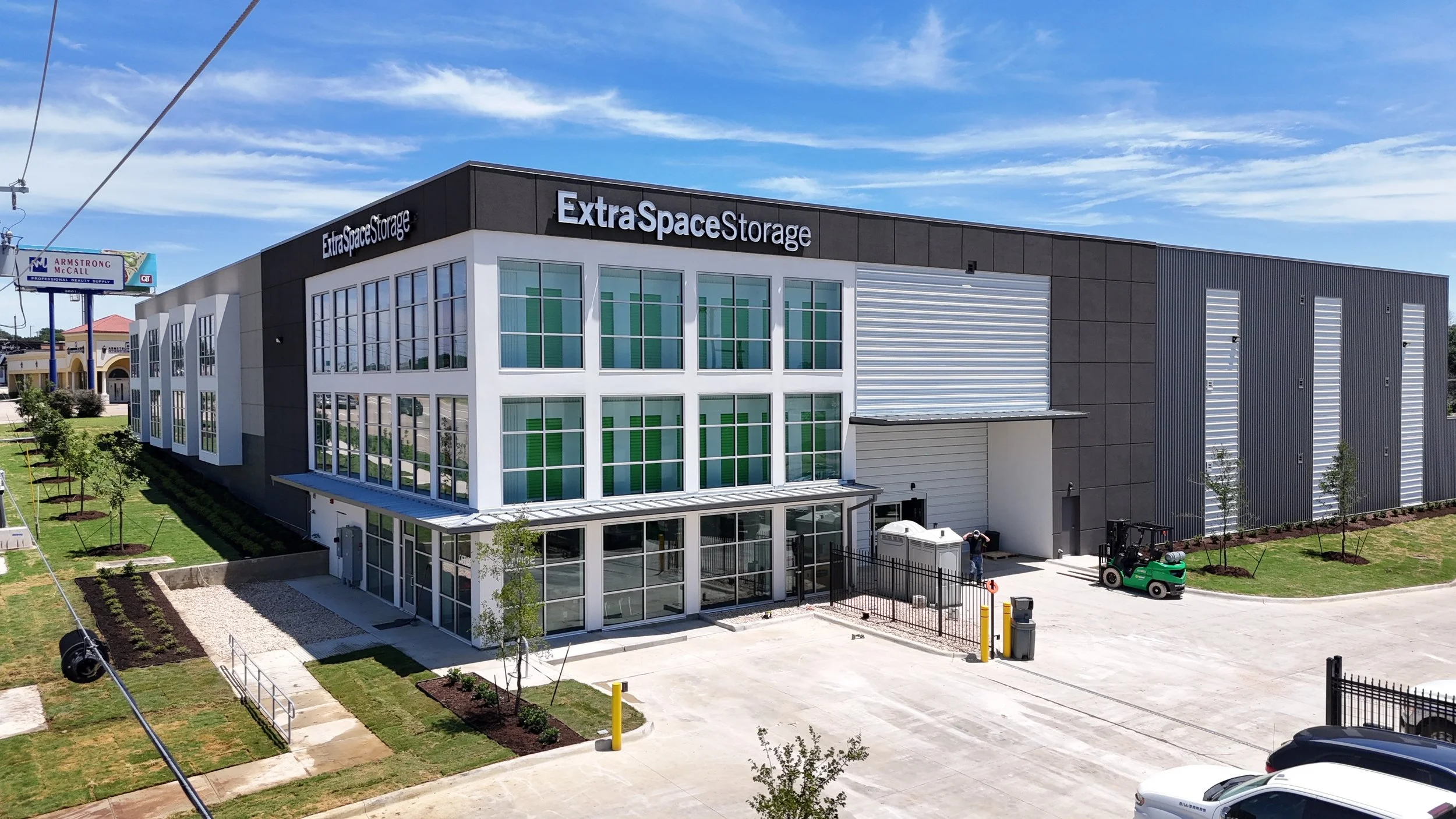 New multi-story storage facility with large glass windows and a black and gray exterior. The sign reads 'ExtraSpaceStorage.' The parking lot is mostly empty with a forklift and some parked cars. Small trees and landscaped areas surround the building.