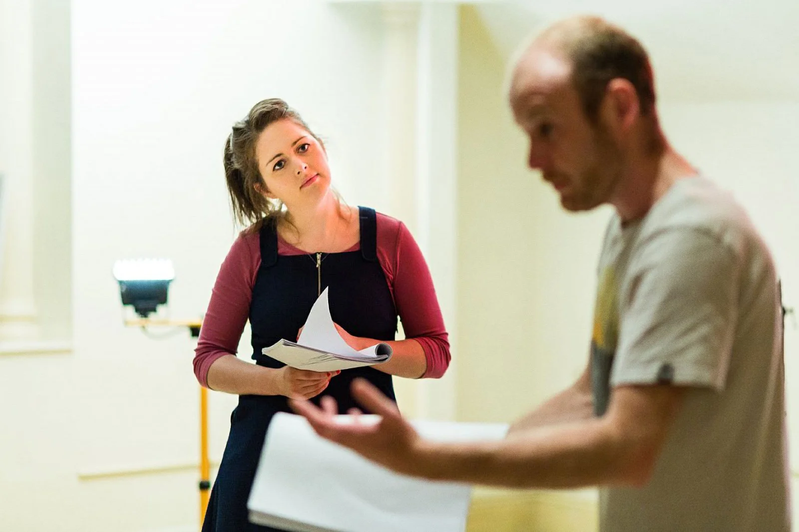 A woman holding papers looks at a man who is in the foreground, reading from a book or a notebook. The background shows a light-colored wall and a light source.