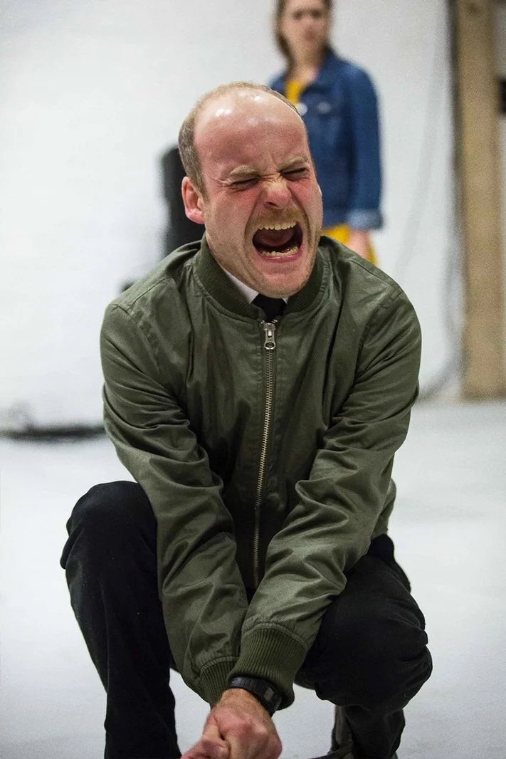 A man in a green jacket is squatting with his hands clenched in fists, appearing to scream or yell with his eyes closed and mouth open. In the background, a woman in a denim jacket is standing and slightly out of focus.