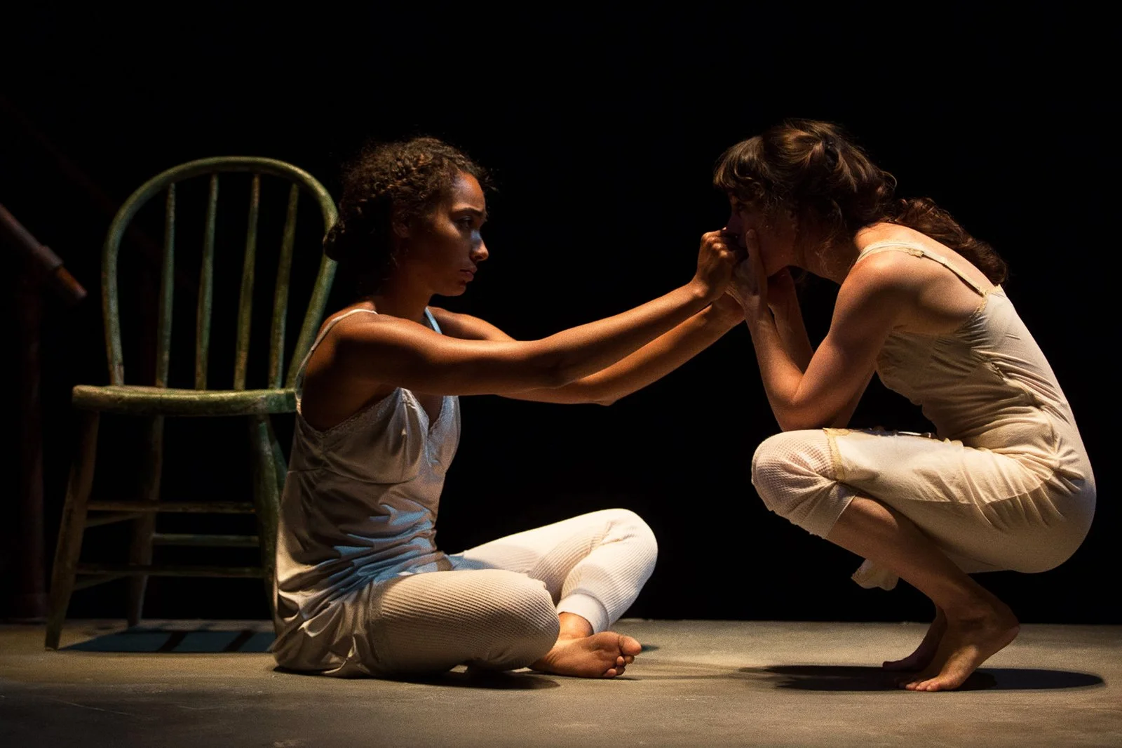 Two women, one sitting on the floor and the other crouching, are engaging in a moment of emotional intimacy, touching faces and holding hands in a dark setting
