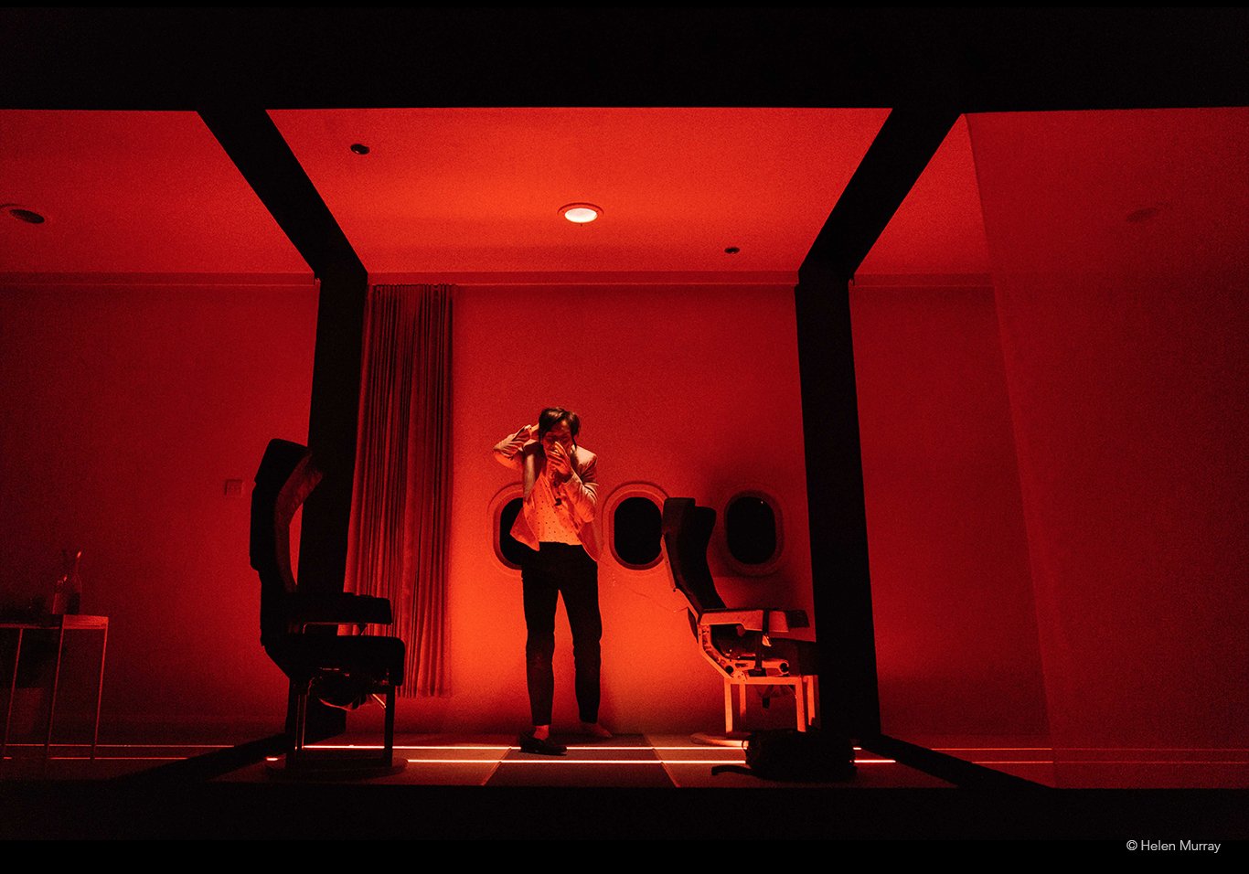 A person standing in a dimly lit room filled with red light, near hair styling chairs and a curtain, with a wall decorated with three small round openings.