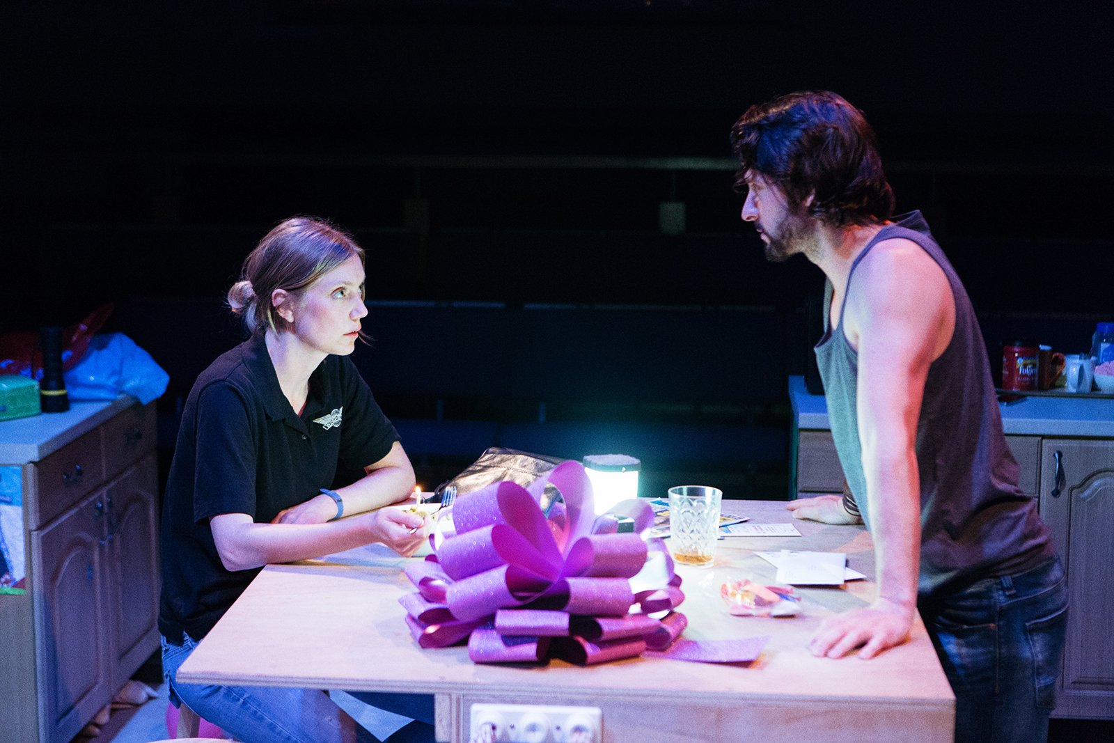 A young woman and a man in a kitchen at night, having a serious conversation across a table with a pink ribbon and wrapping paper.