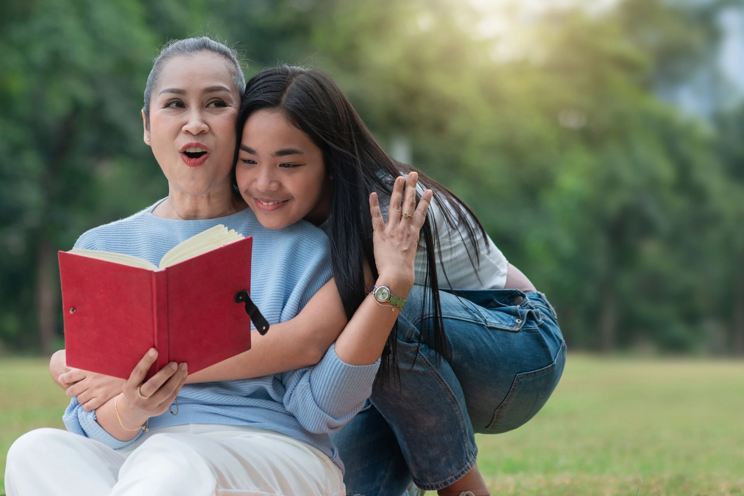 A woman and a girl reading a book together outdoors in a park, with the woman surprised and the girl smiling.