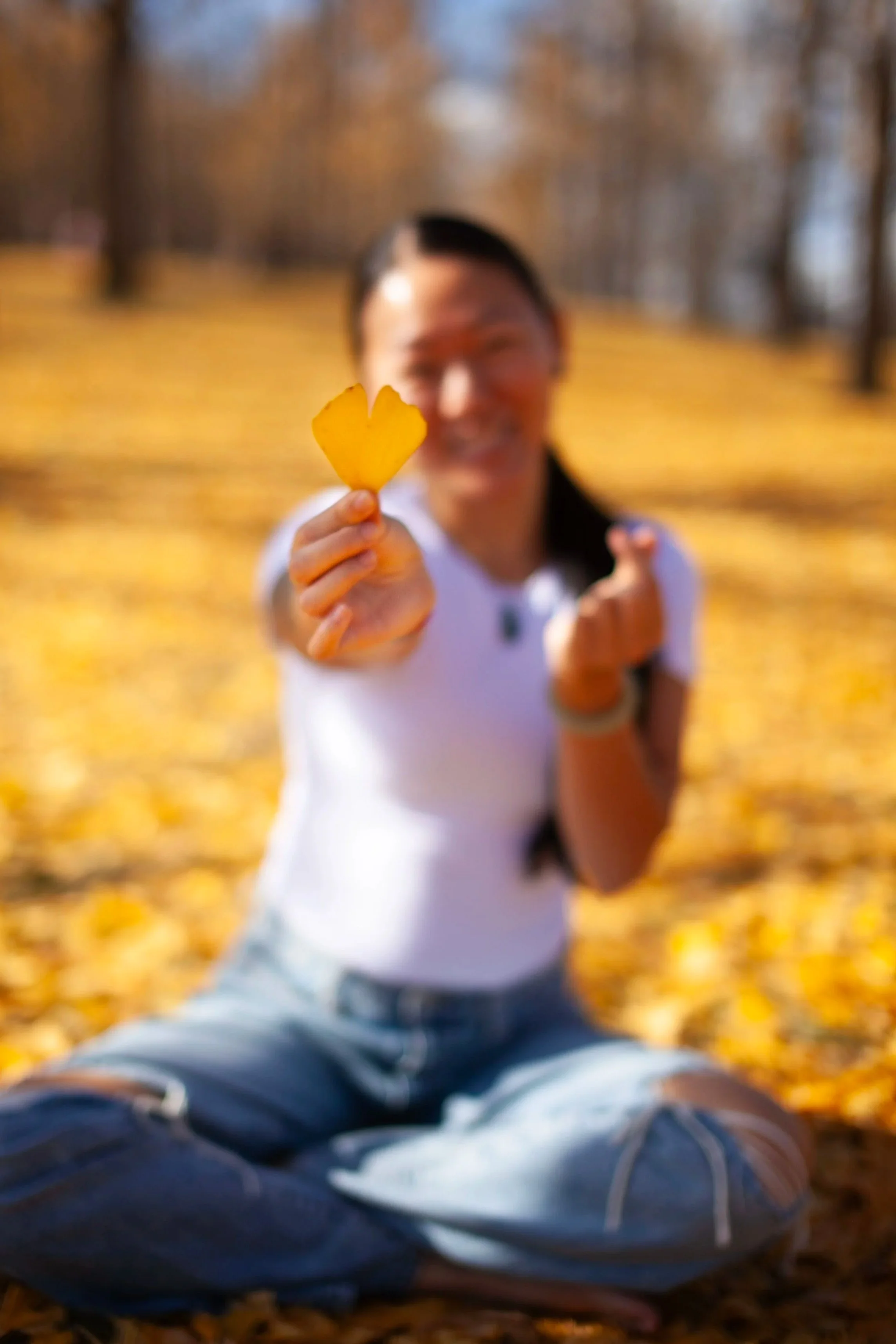 Person sitting on the ground in a park with fallen yellow leaves, holding a yellow leaf close to the camera, smiling.