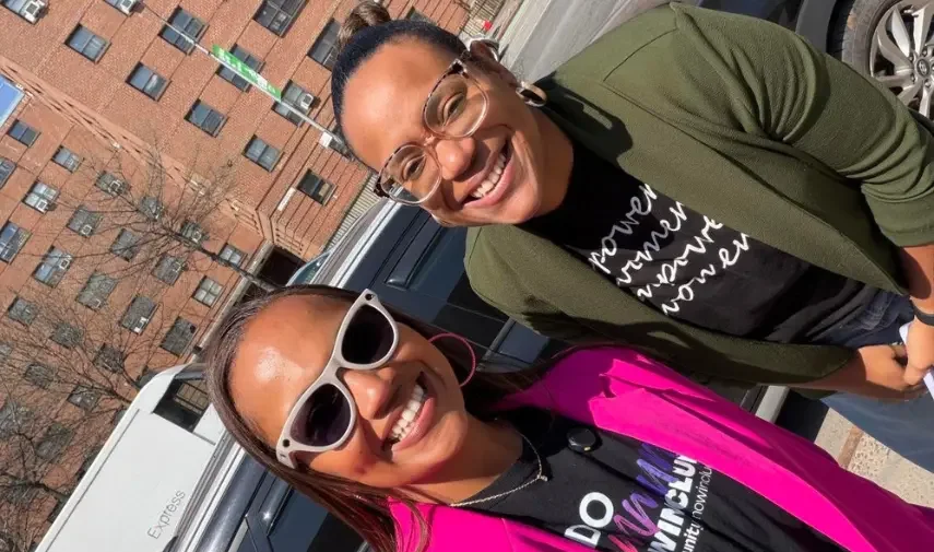 Two women smiling outdoors in front of a vehicle and brick buildings, wearing branded community event shirts