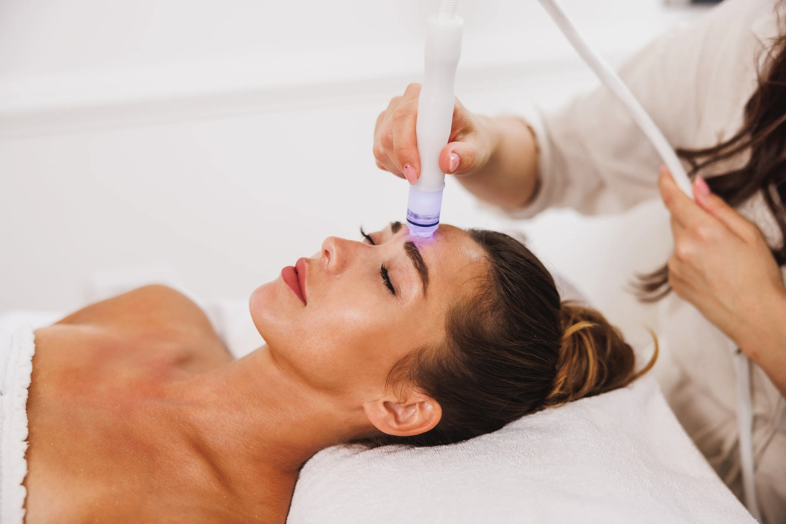 White woman lays with her eyes closed while an aesthetician places a hydrofacial treatment device on her forehead.