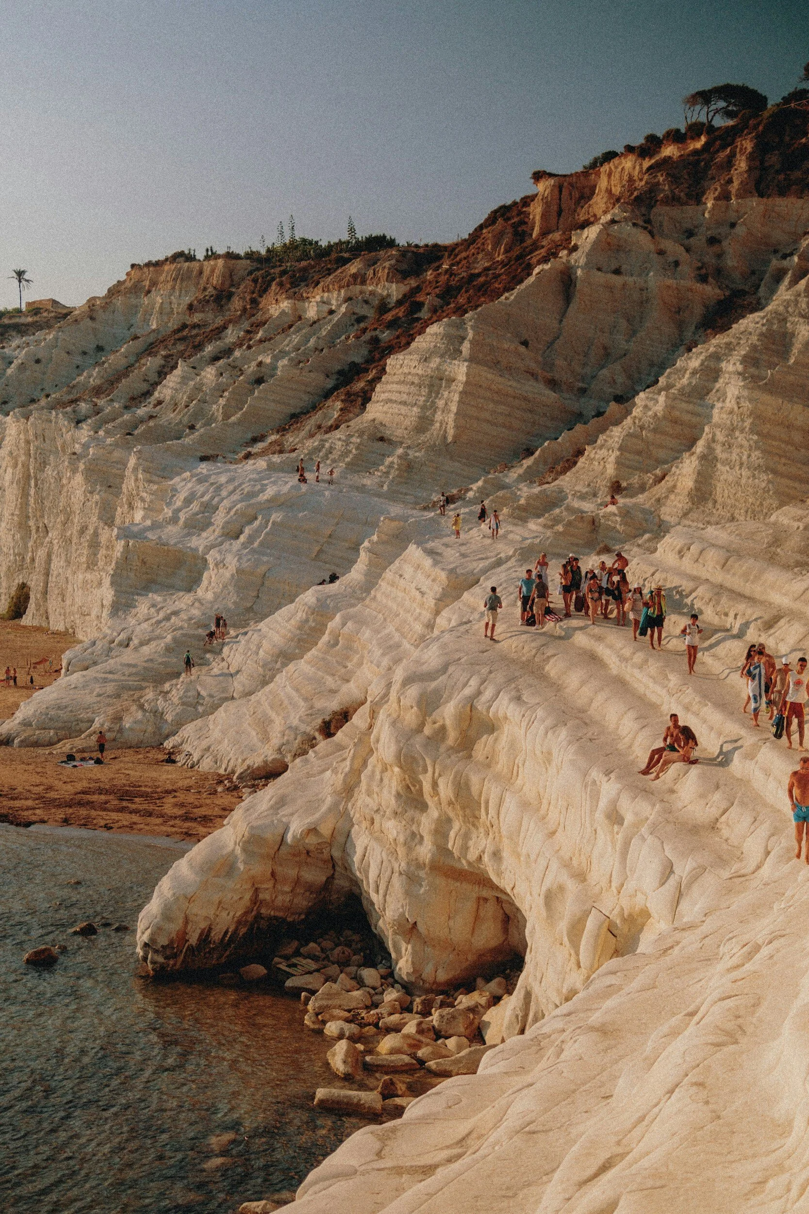 People walking and sitting on white limestone cliffs by the ocean at sunset.