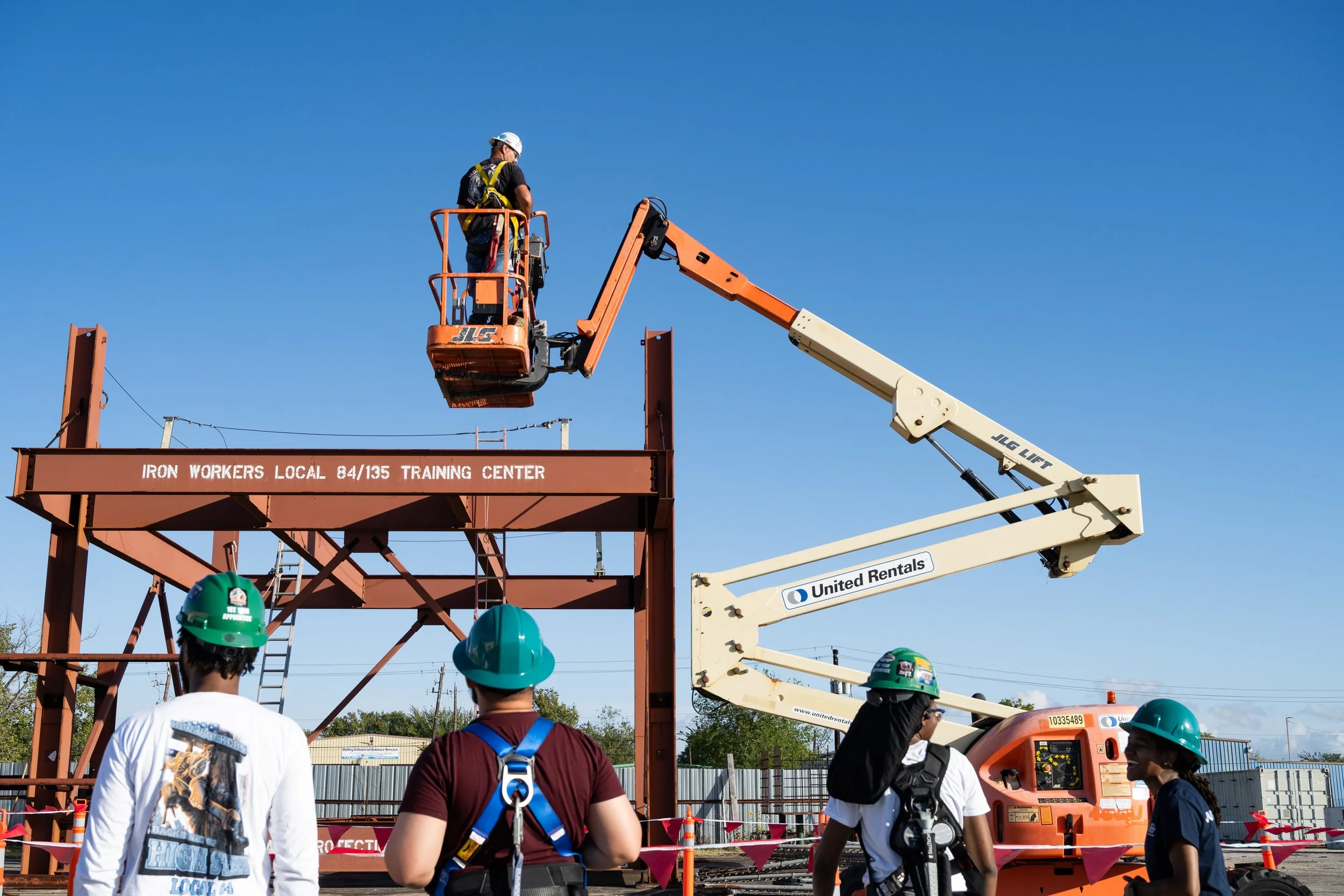 Ironworkers local 84/135, four apprentices stand talking as an instructor and apprentice go up in the arial lift during training