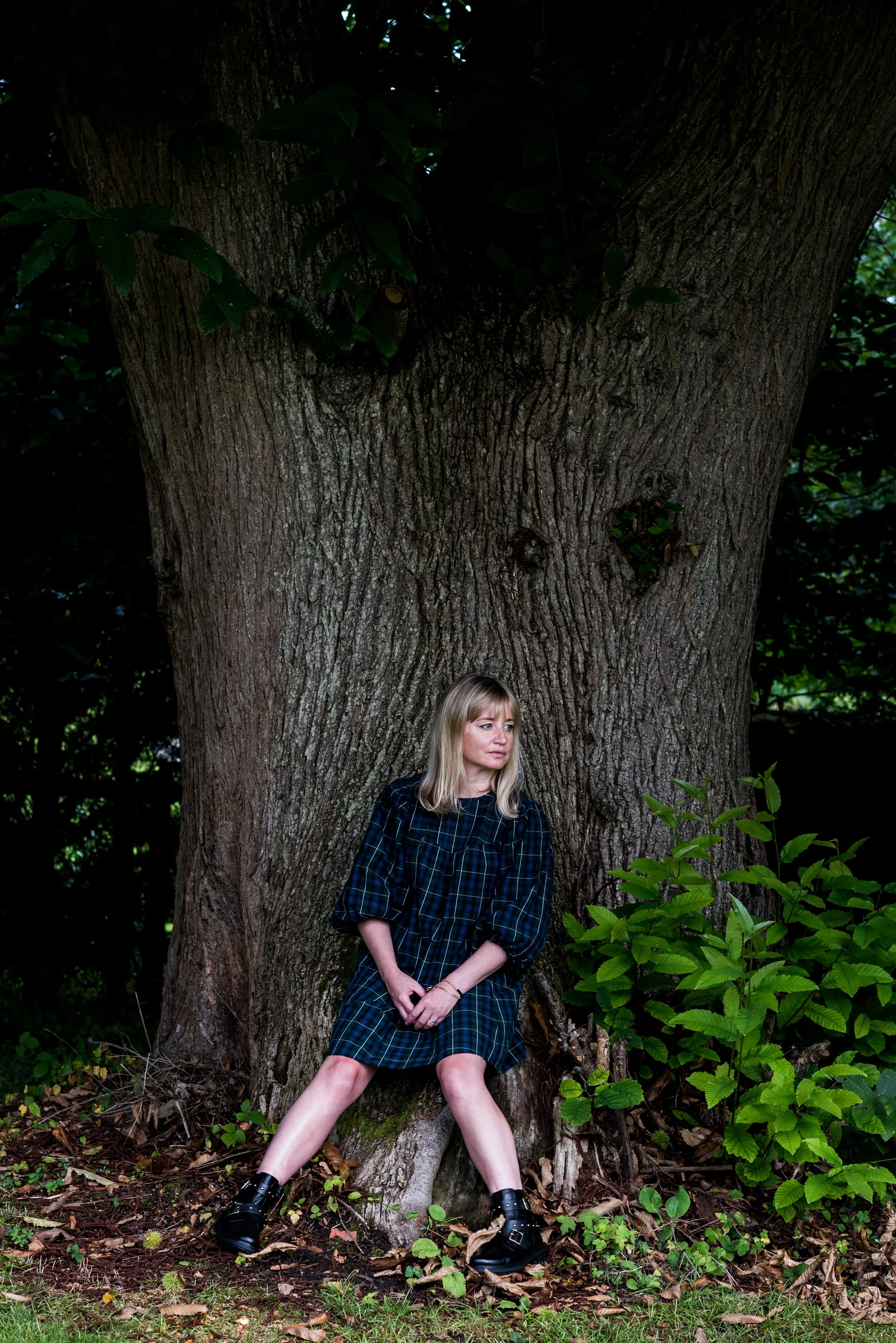 A woman with blonde hair wearing a blue and black plaid dress, black boots, standing in front of a large tree in a forested area. She is posing with her legs apart, hands clasped in front, with greenery and leaves around her.