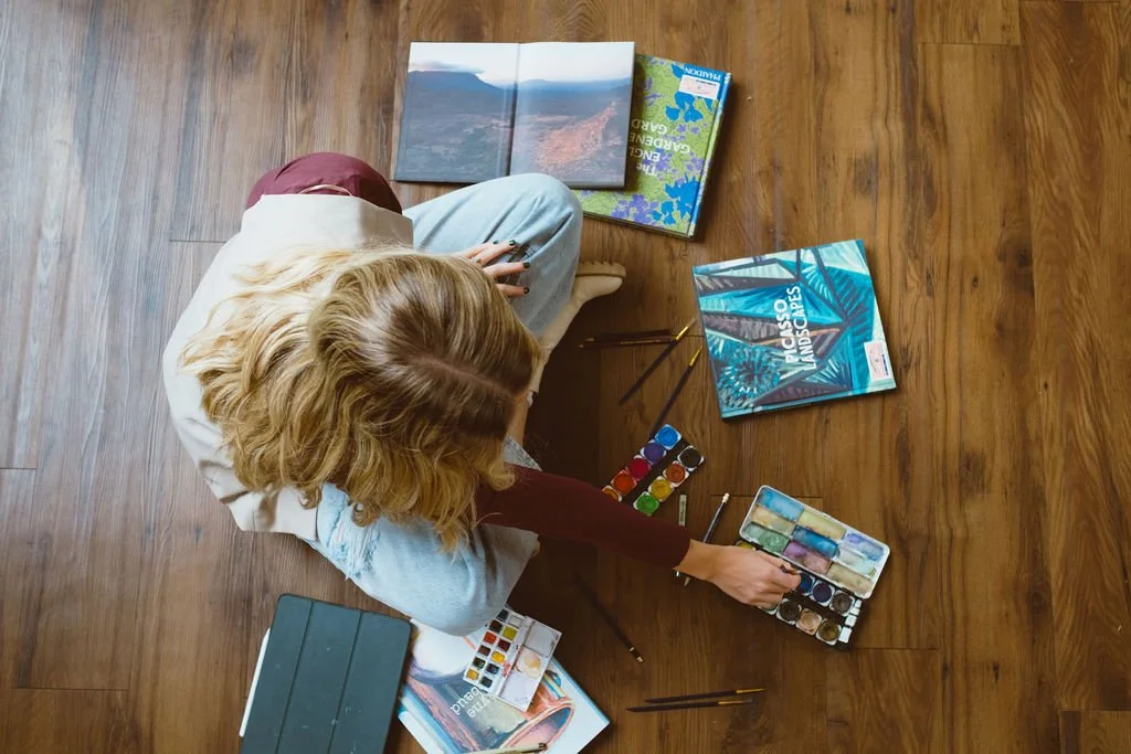 A person sitting on a wooden floor surrounded by opened books, watercolor paint sets, brushes, and a tablet, engaged in painting or drawing.