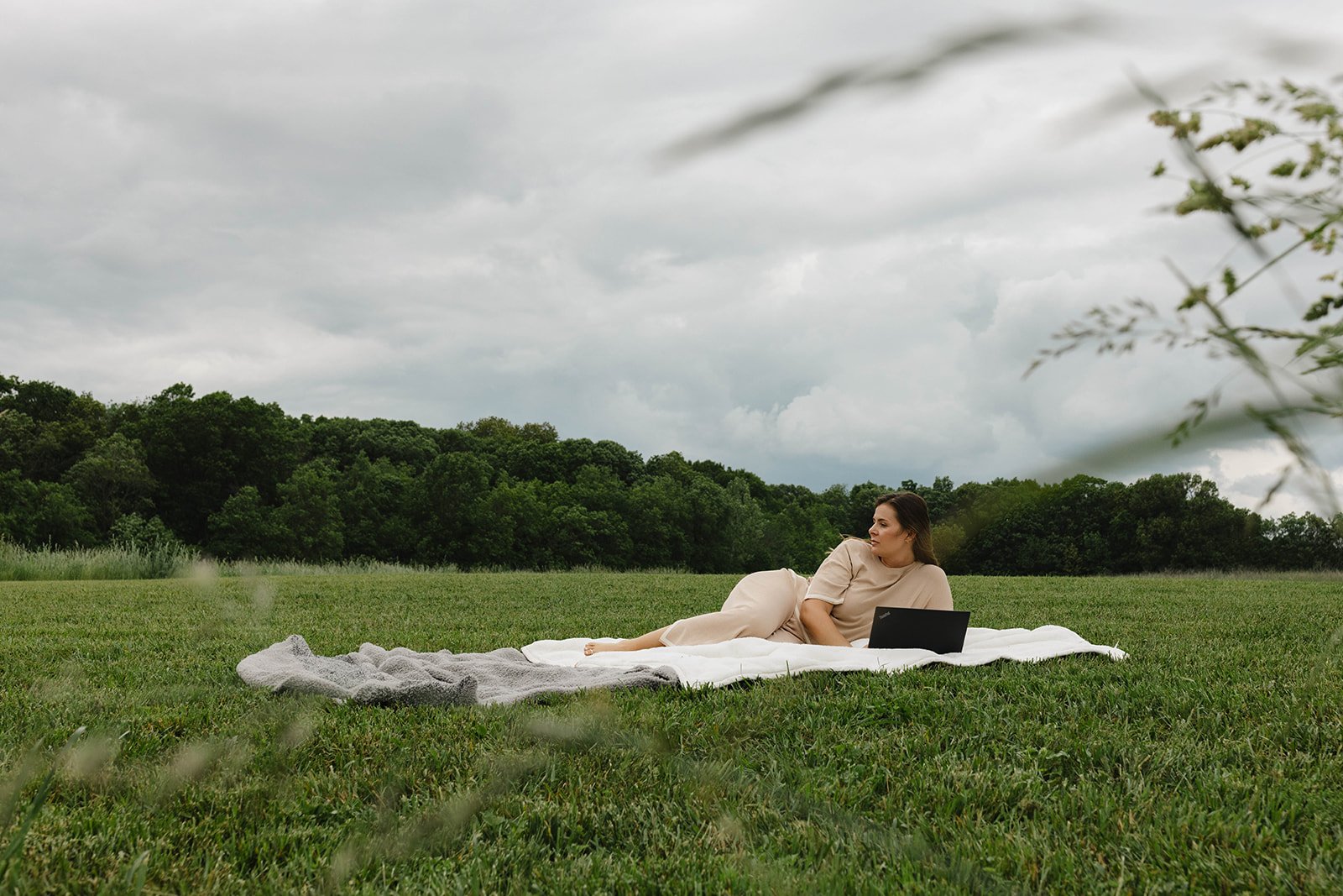 Kerri M. Roberts, keynote speaker and podcast host, lying on a white blanket in a grassy field with a laptop nearby, under a cloudy sky, with green trees in the background.