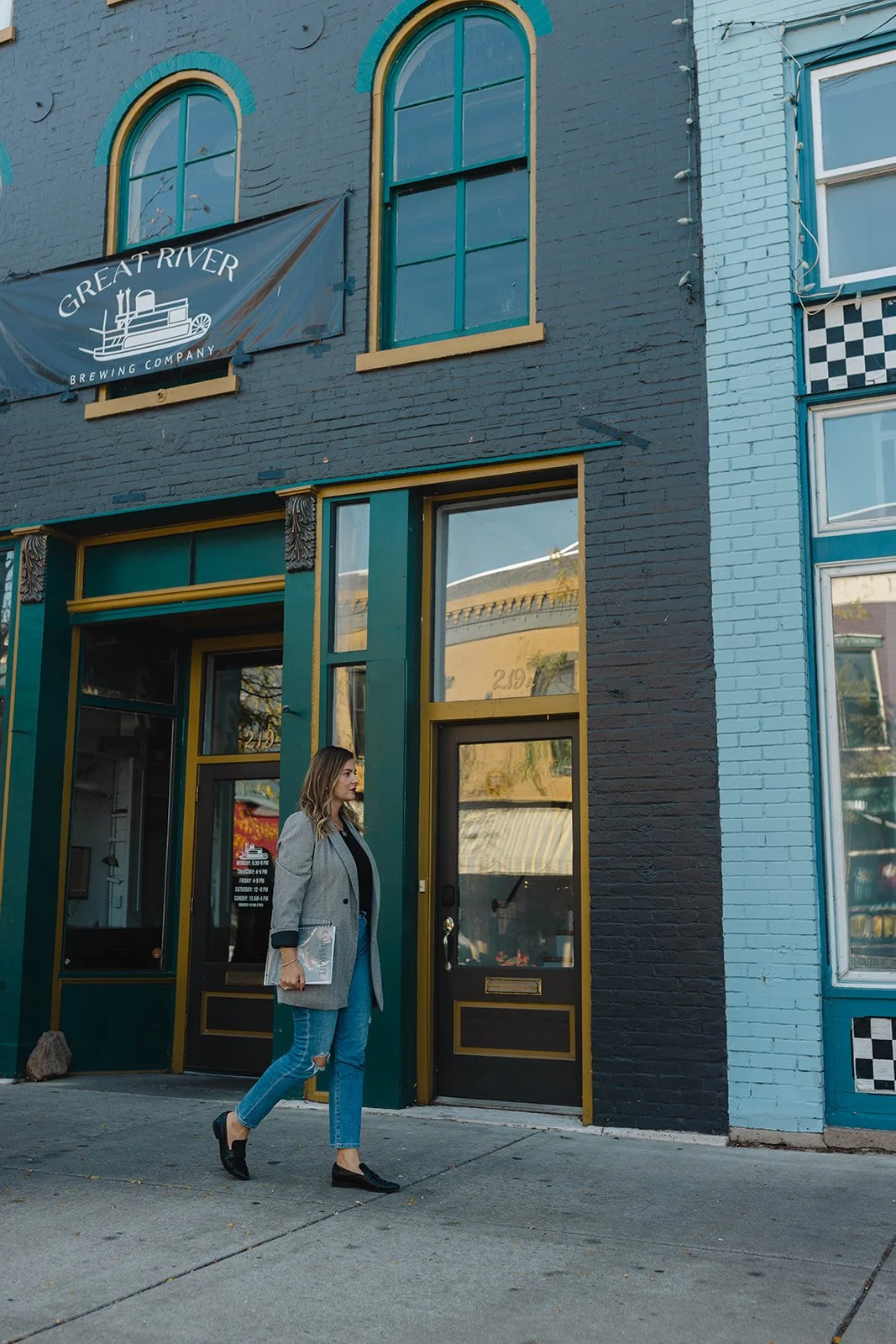 Kerri M. Roberts, keynote speaker and podcast host, walking past a colorful building with large windows and a sign for Great River Brewing Company.