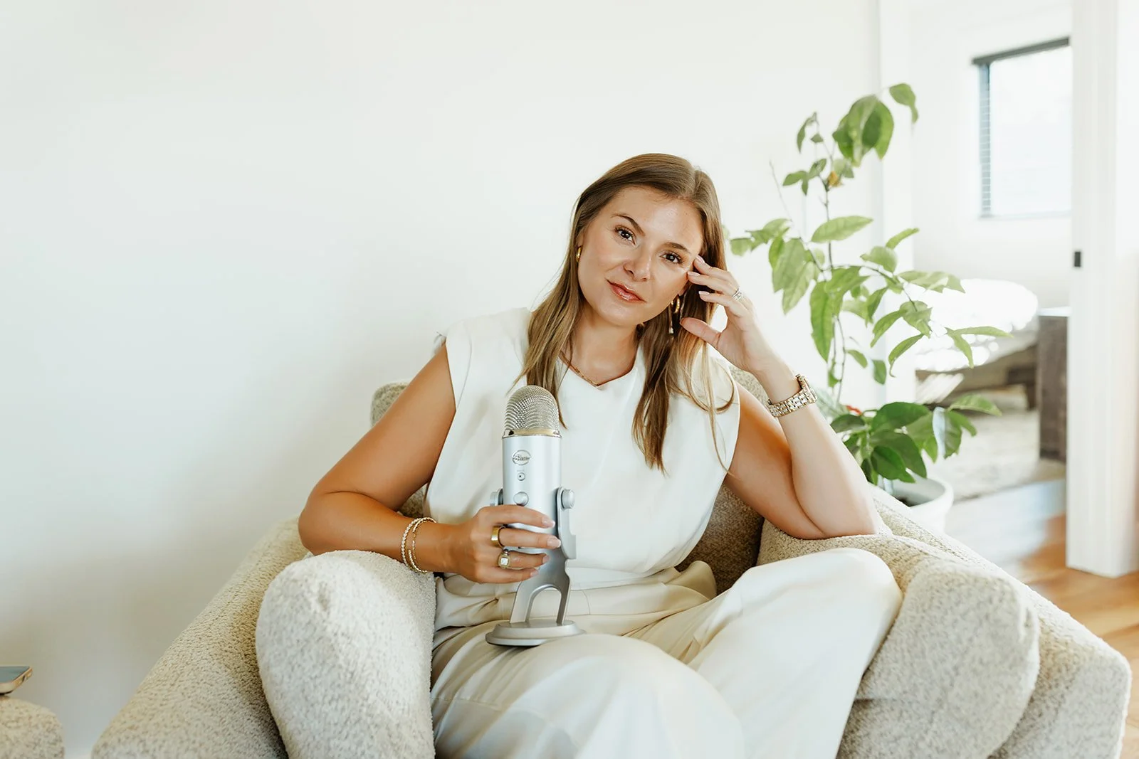 Kerri M. Roberts, keynote speaker and podcast host, sitting on a beige armchair, holding a silver microphone, and looking at the camera in a bright room with a green plant and a bed in the background.