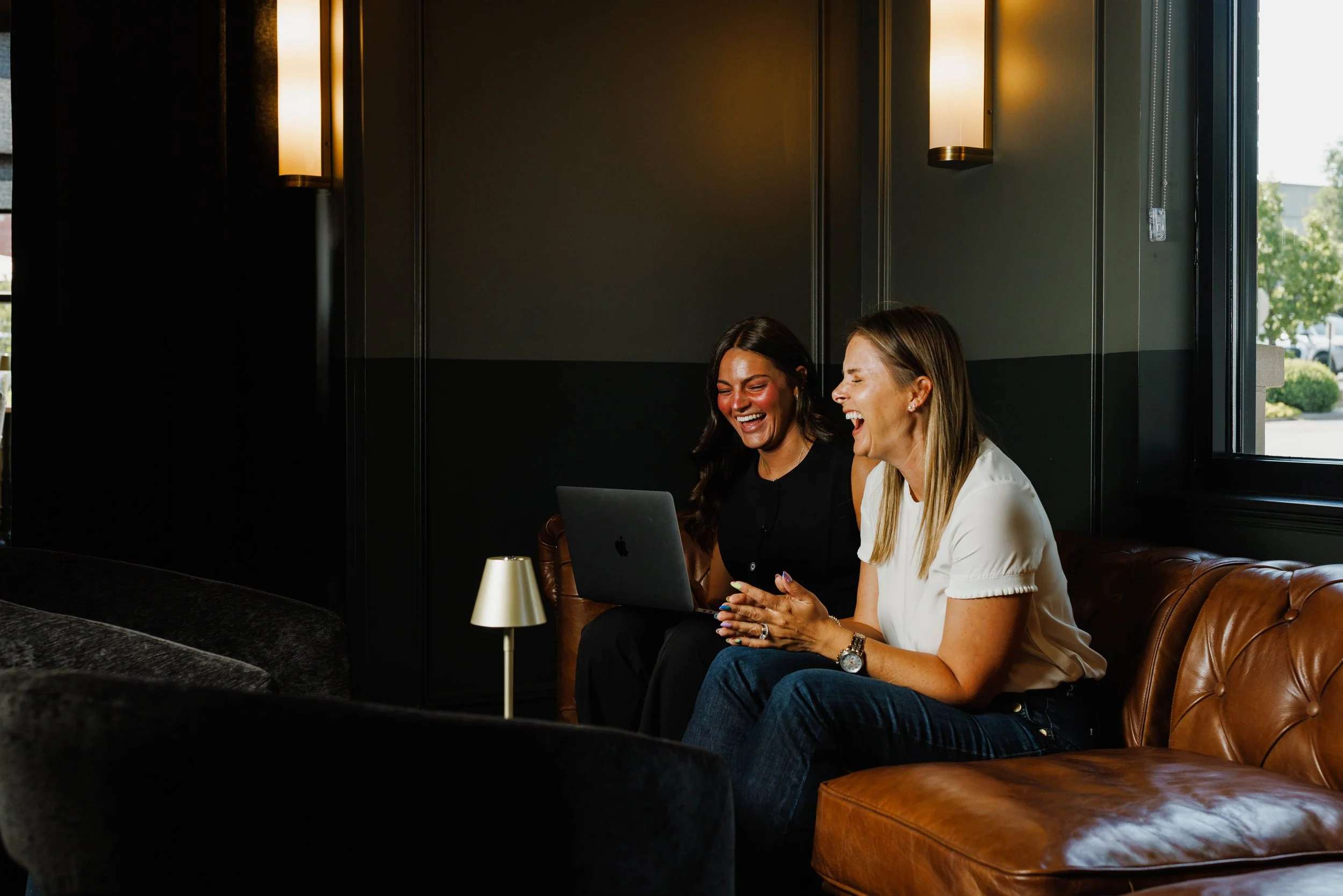 Two women sitting on a leather couch, laughing and looking at a laptop in a dimly lit room with a window showing outdoor greenery.