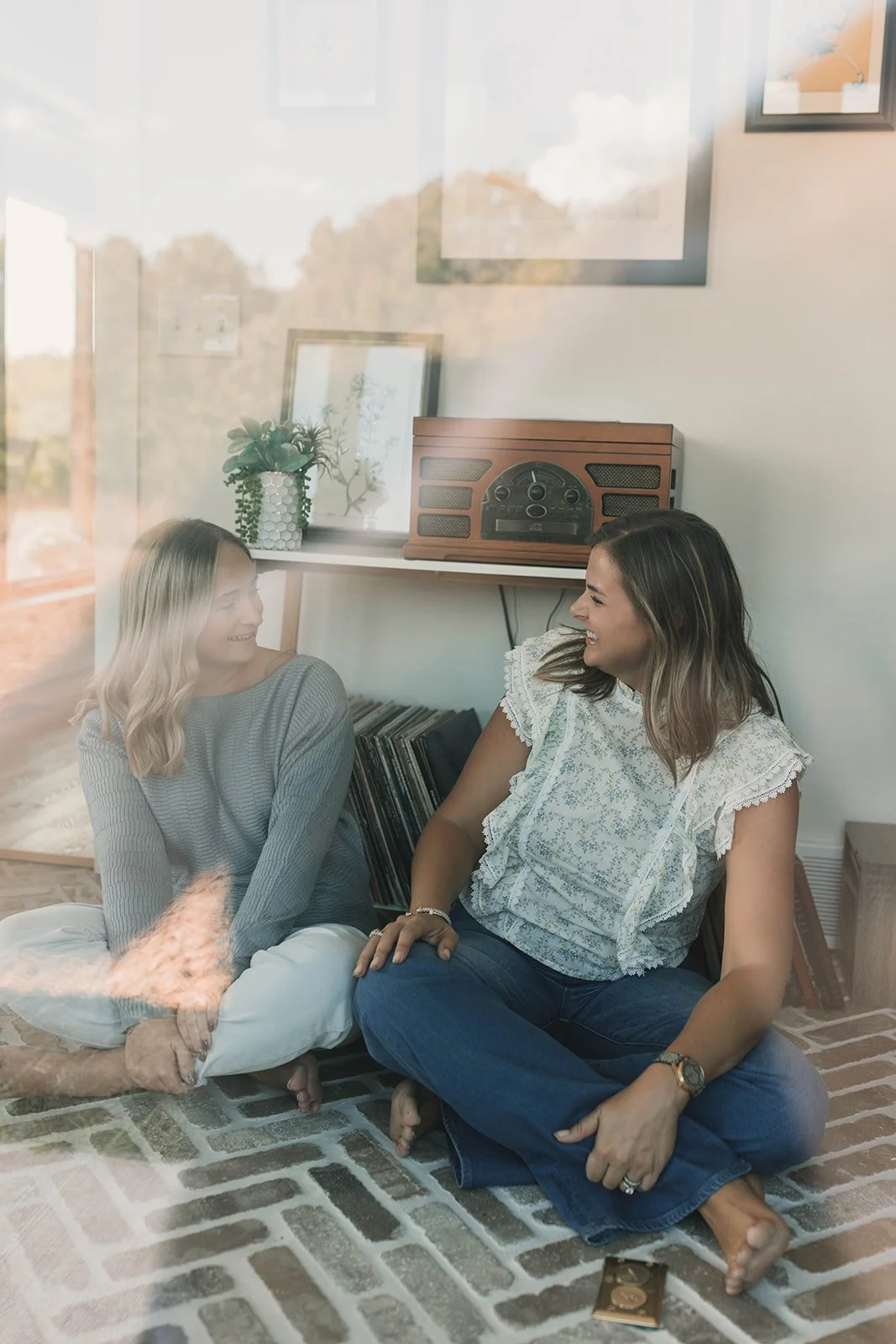 Kerri M. Roberts, keynote speaker and podcast host, with a member of her team, sitting on the floor and smiling at each other in a cozy living room with a retro radio, vinyl records, and framed pictures on the wall.