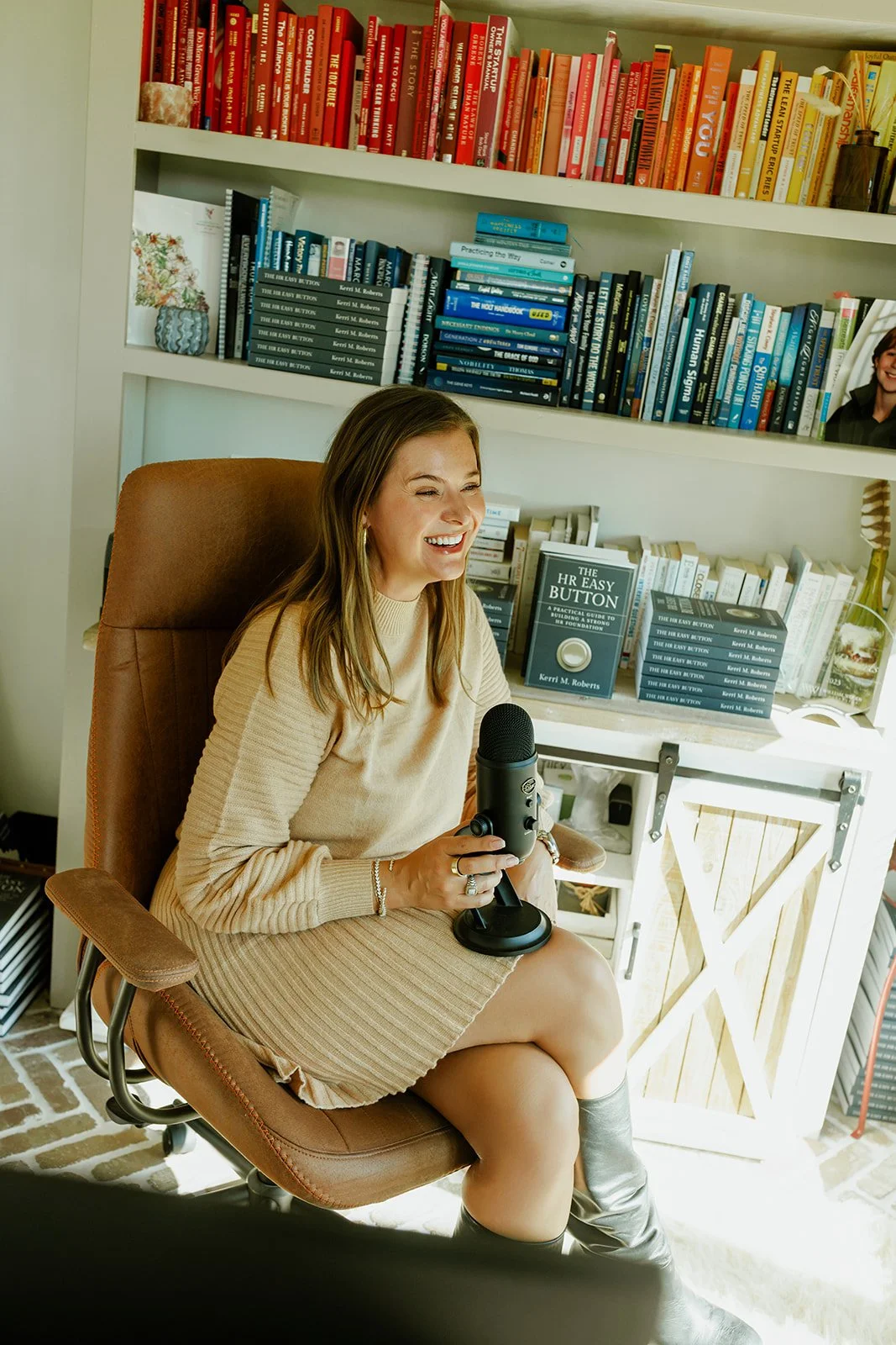 Kerri M. Roberts, keynote speaker and podcast host sitting in a brown office chair, holding a microphone, smiling, in a room with bookshelves filled with colorful books, including a collection of books titled 'The HR Easy Button' by Kerri M. Roberts.