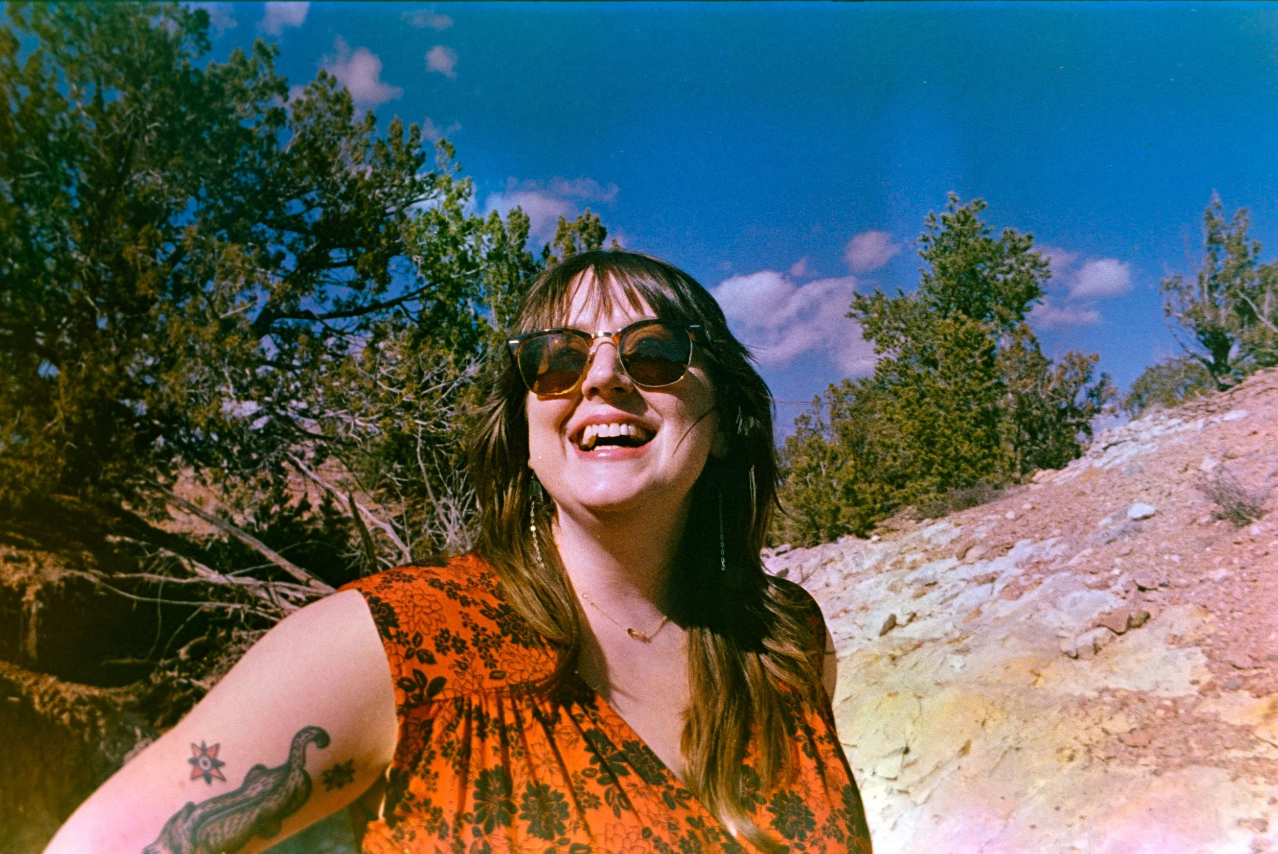 A smiling woman with sunglasses, wearing an orange floral dress, standing outdoors in a rocky landscape with trees and a blue sky with clouds.