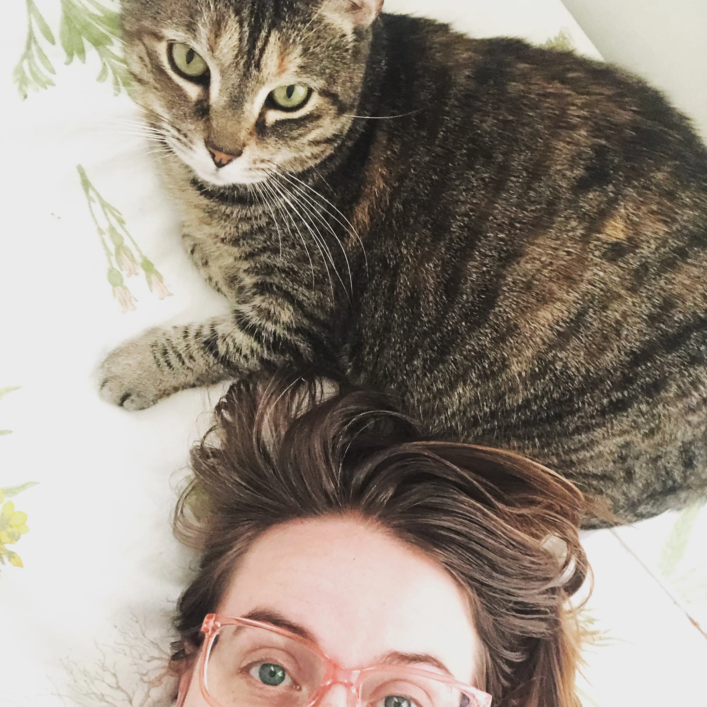 A woman with reddish-brown hair and pink glasses lying on a bed with a gray tabby cat beside her, both looking up at the camera. The bed has white sheets with green floral patterns.