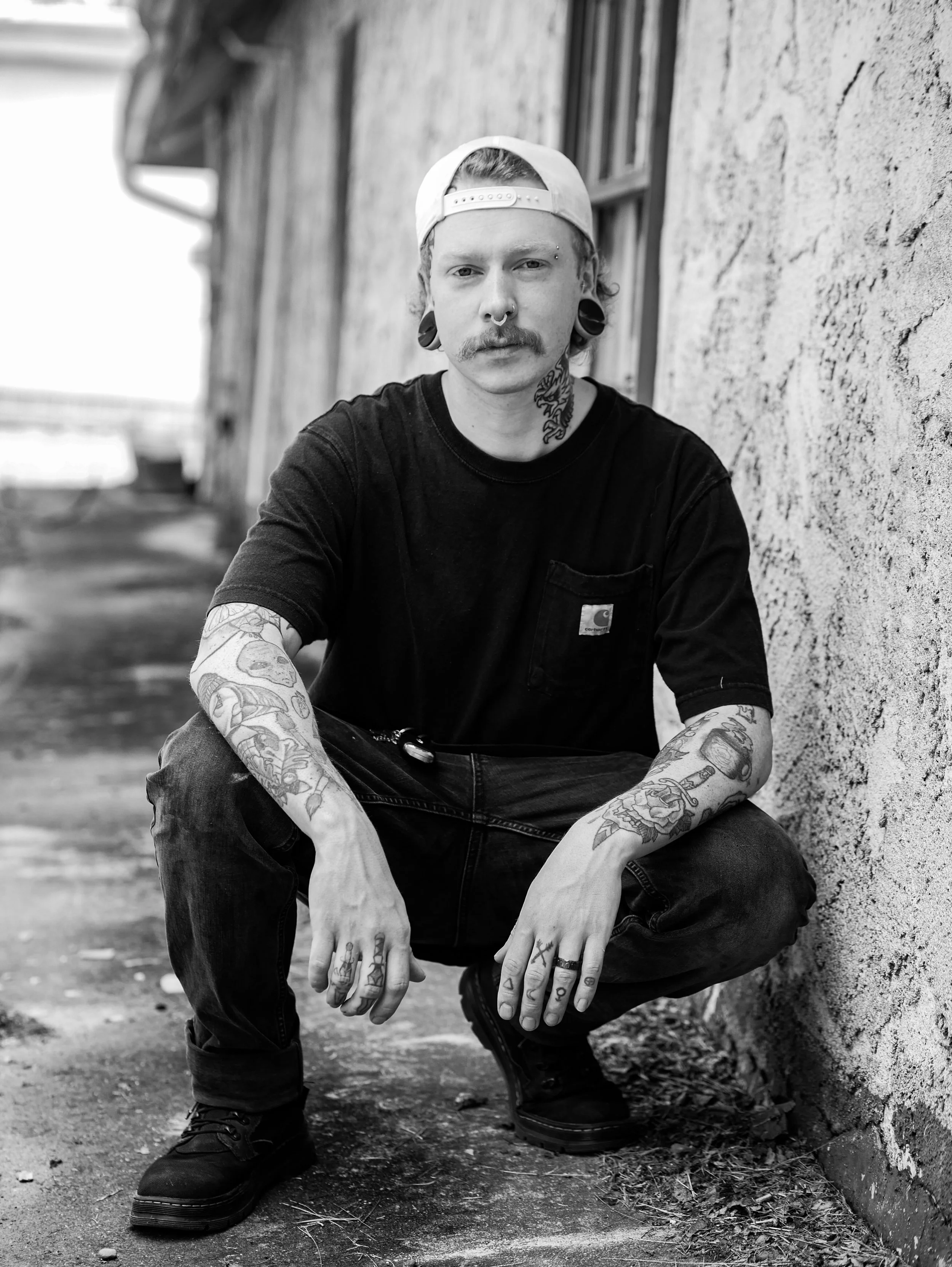 A man crouching next to a textured wall with visible tattoos on his arms and neck, wearing a backwards cap, black t-shirt, and black boots.