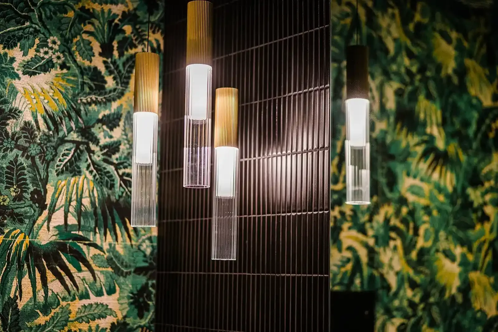 Elegant brass and fluted glass pendant lighting hanging in front of a vibrant tropical leaf wallpaper at Smile Bar in Calgary.