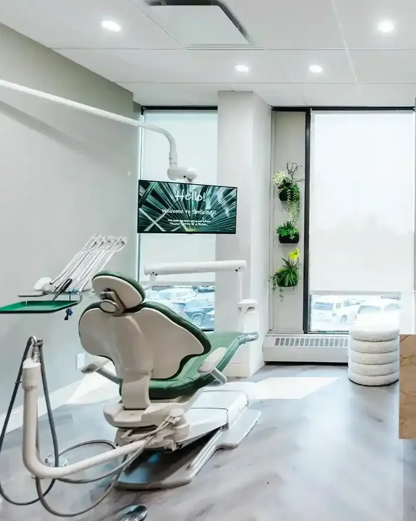 A modern dental treatment room featuring a green patient chair, a welcome screen, and a large window at Smile Bar in Calgary.