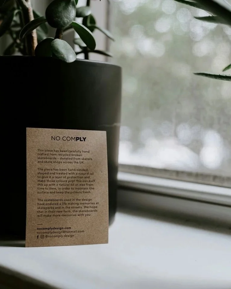 A black plant pot with a green plant next to a window, and a brown Kraft care card with black text about a piece made from recycled skateboard parts.