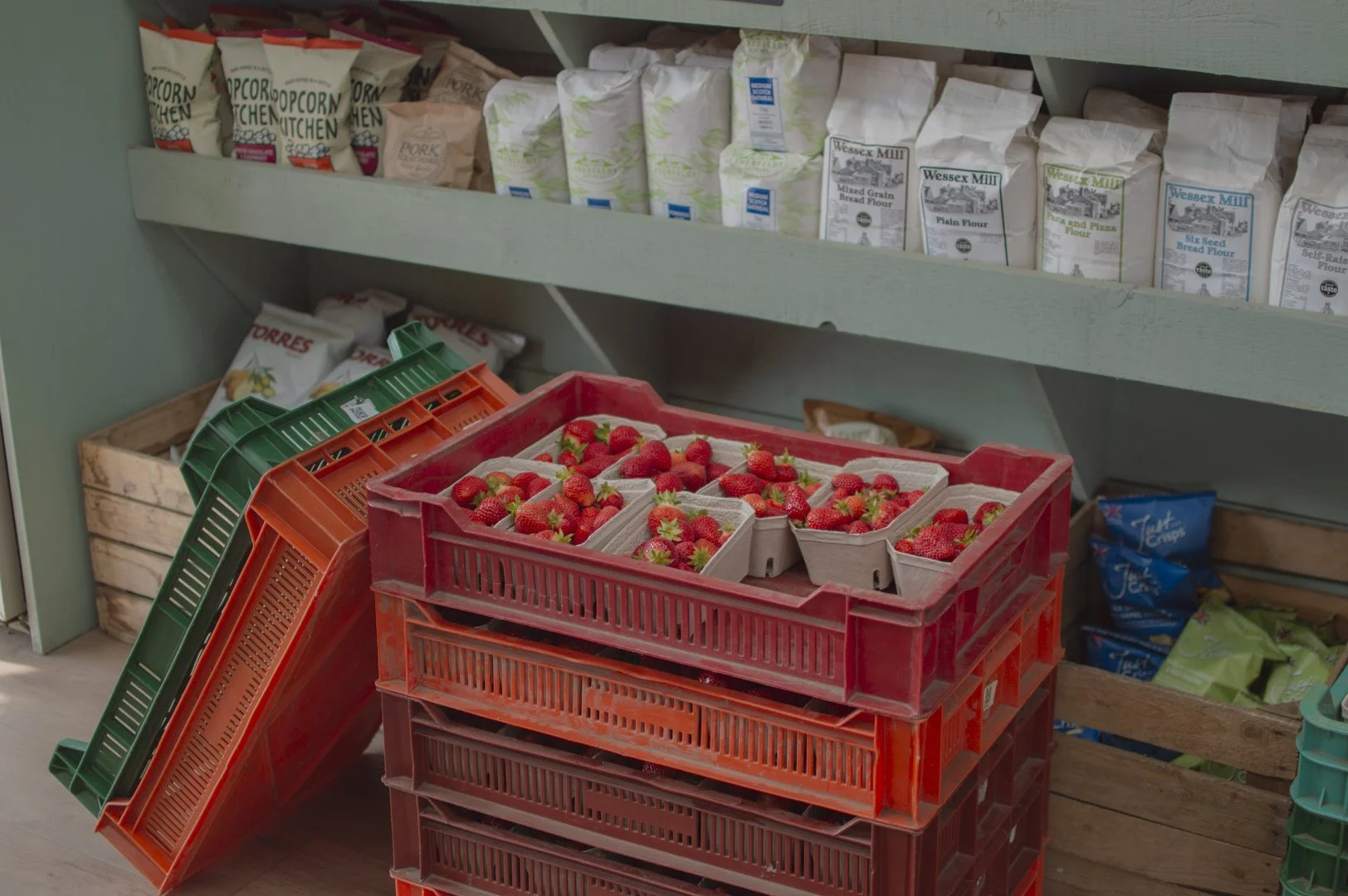 Red plastic crates filled with fresh strawberries in a market or grocery store. Shelves in the background hold various packaged flours.