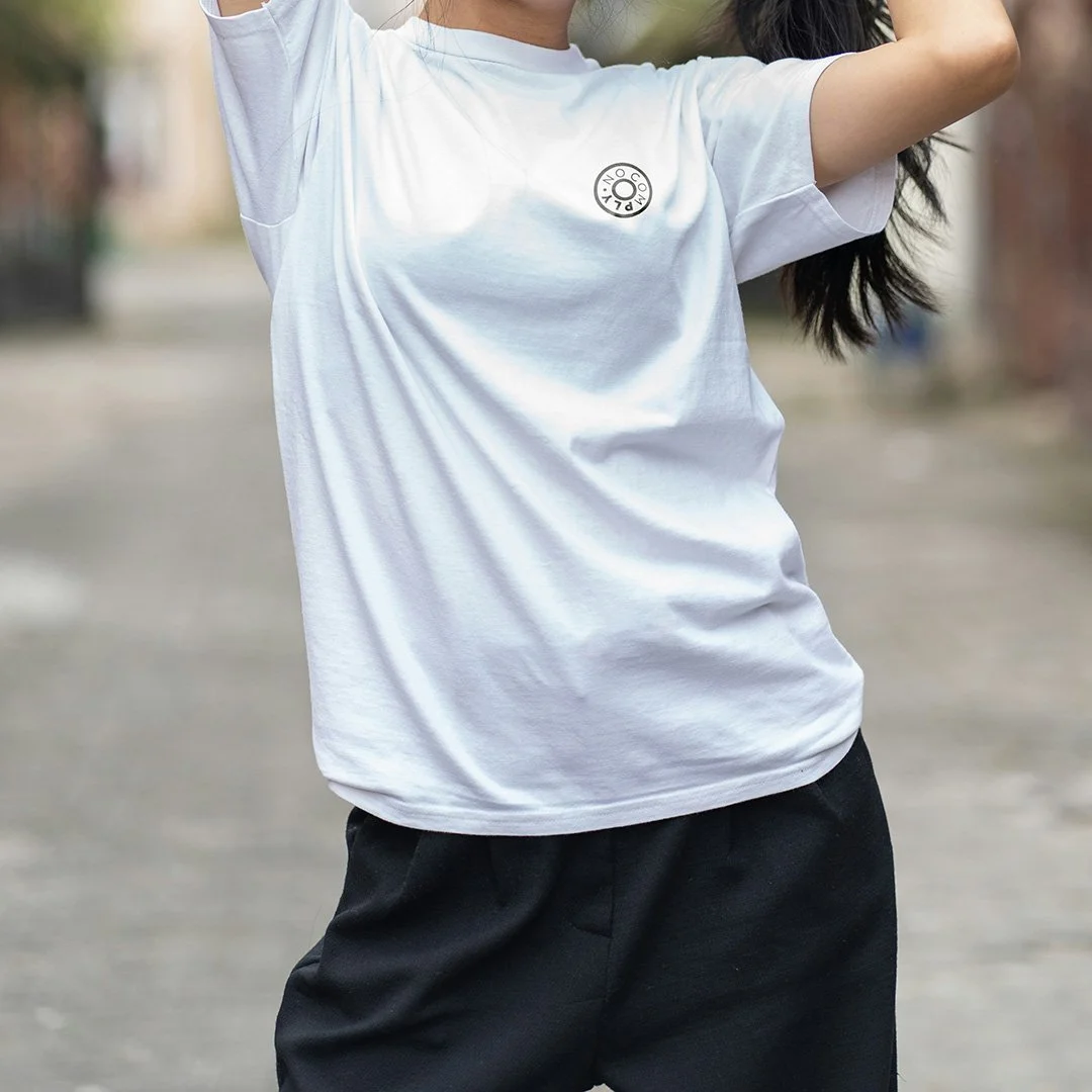 Person wearing a white T-shirt with a small no comply bearing logo on the chest, standing outdoors with one arm raised.