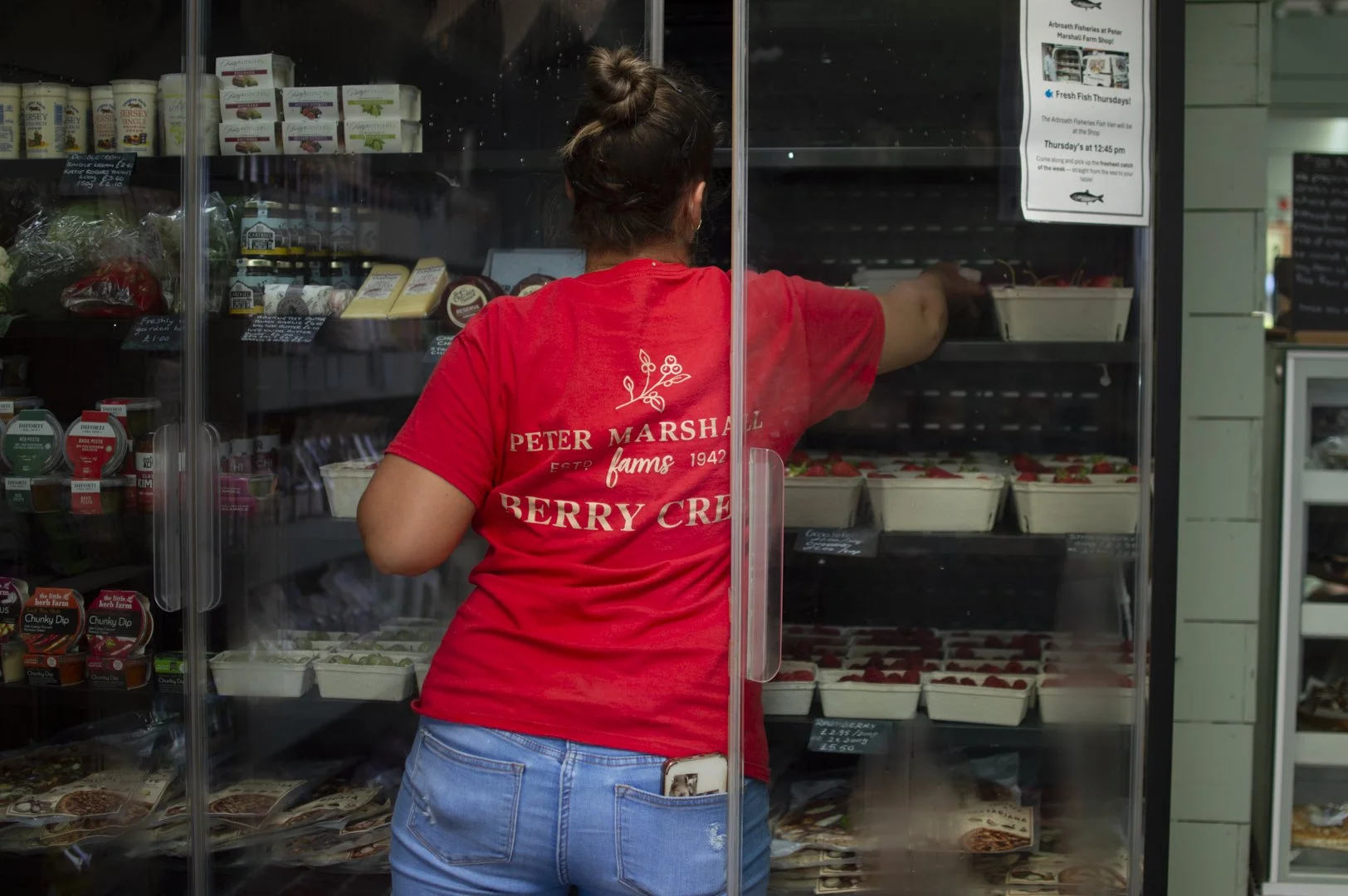 A woman with a bun hairstyle wearing a red T-shirt and blue jeans placing strawberries into a container in a glass display case at a grocery or market store.