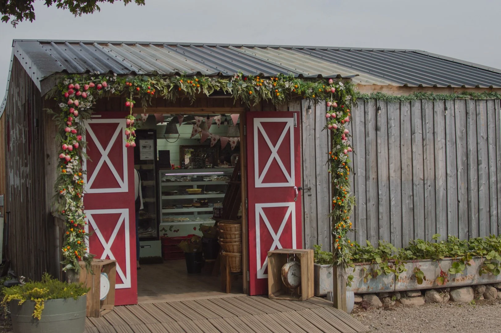 A small rustic shop with red barn-style doors decorated with pink and orange flowers and greenery, located in a wooden shed with a corrugated metal roof and surrounded by planters with greenery.