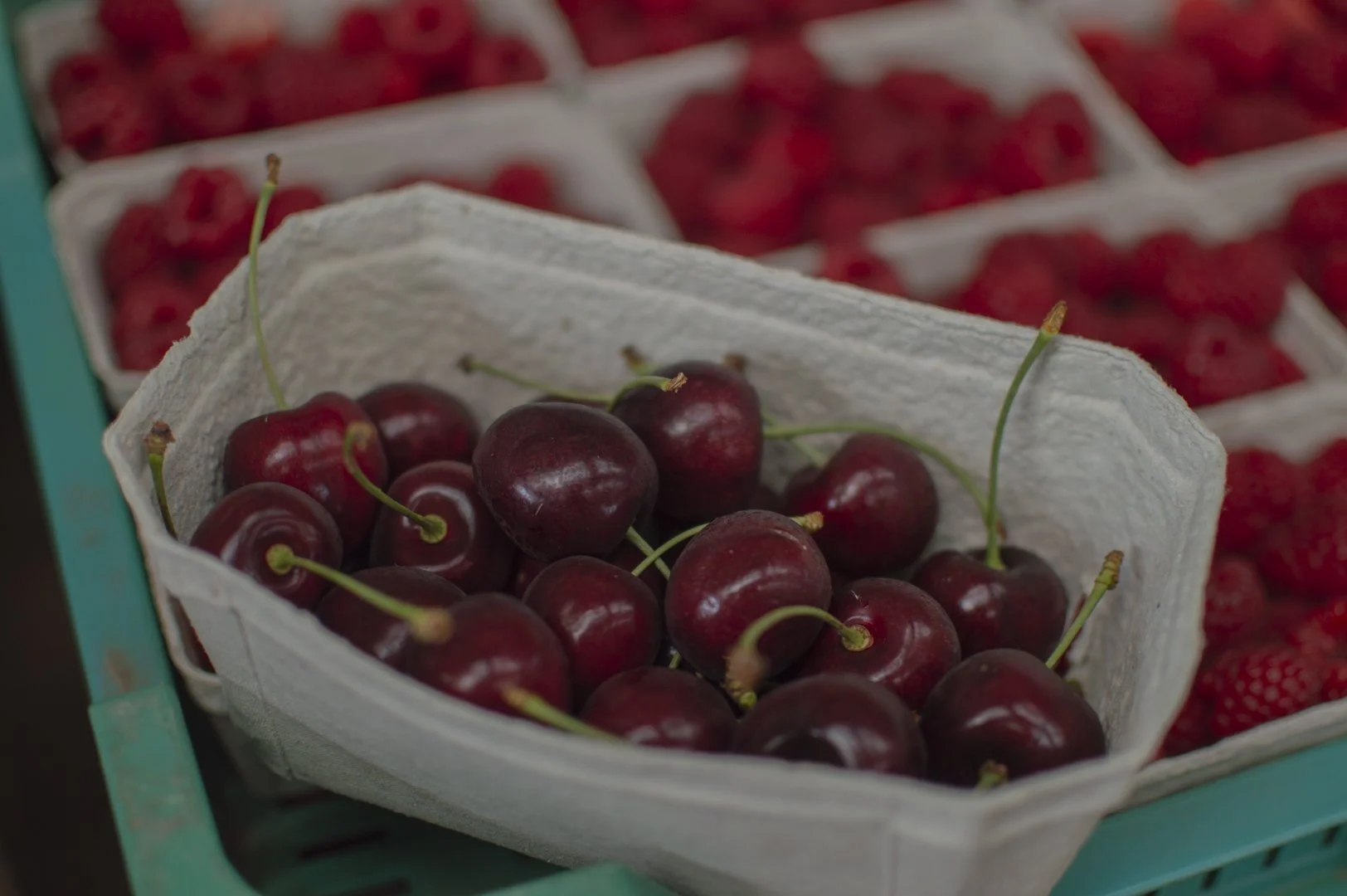 A box of fresh red cherries in a white paper container, with more cherries and strawberries visible in the background.