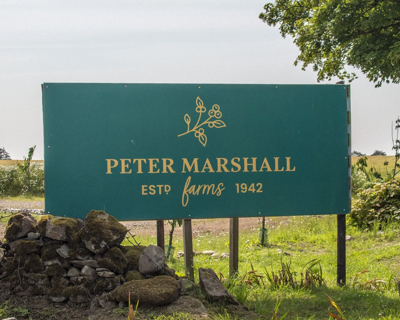 Green sign reads 'Peter Marshall Farms EST 1942' with a leaf illustration above the text, standing on a grassy area near rocks and trees.