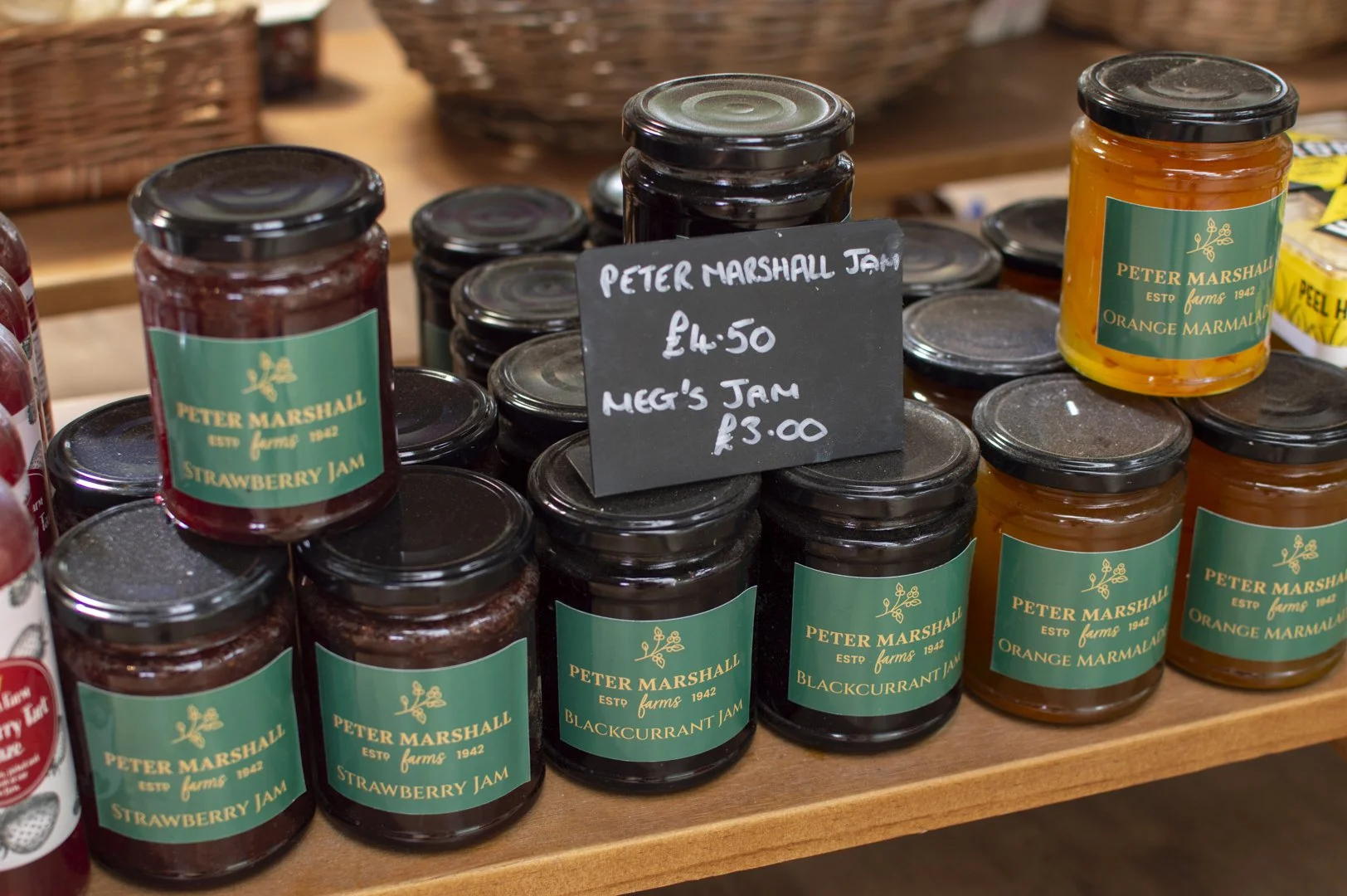 Rows of jars of strawberry and orange marmalade with green labels, displayed on a wooden shelf at a market.