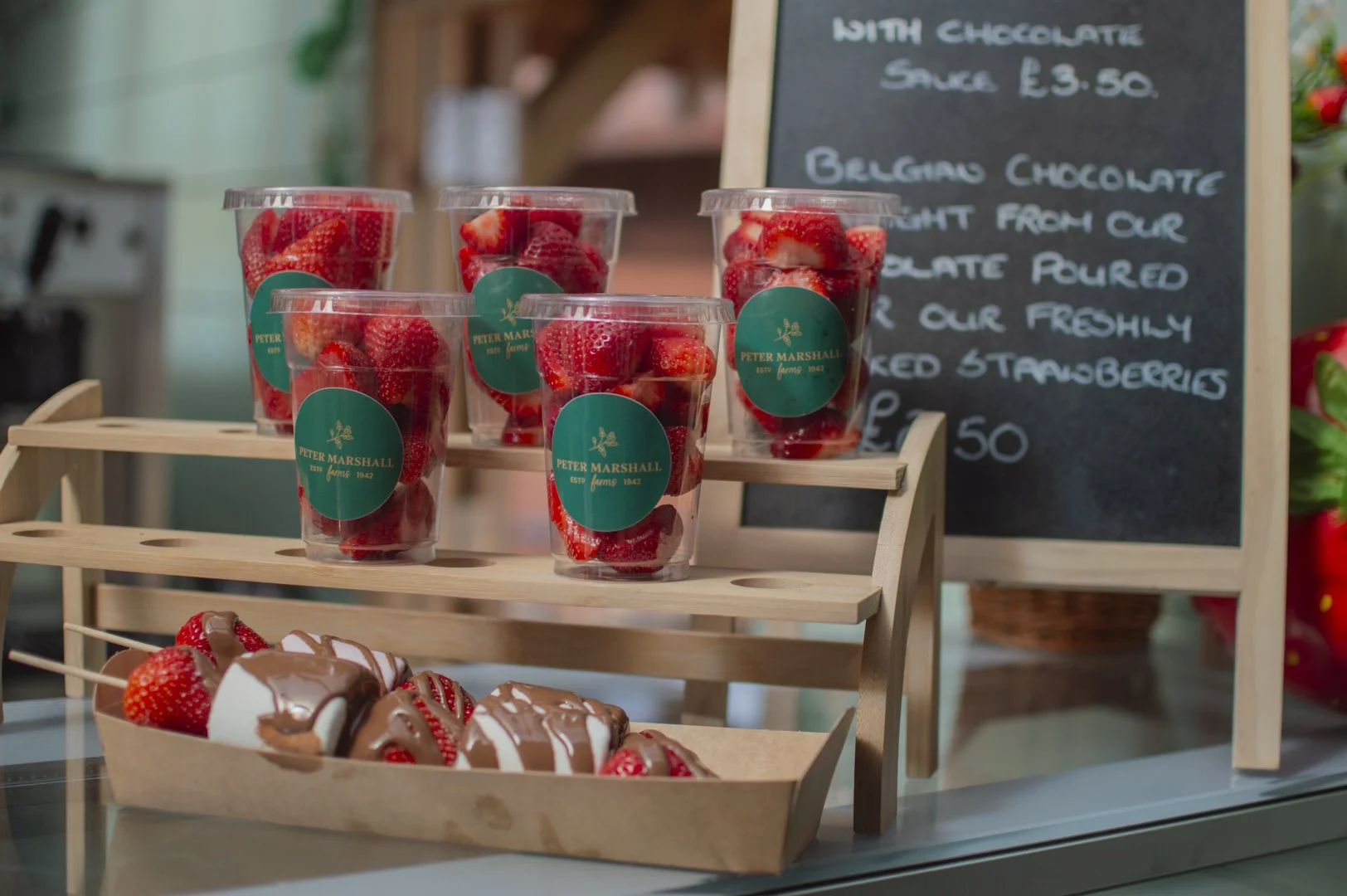 Clear plastic cups filled with fresh strawberries on a small wooden display stand, with a chalkboard sign in the background advertising chocolates and strawberries.