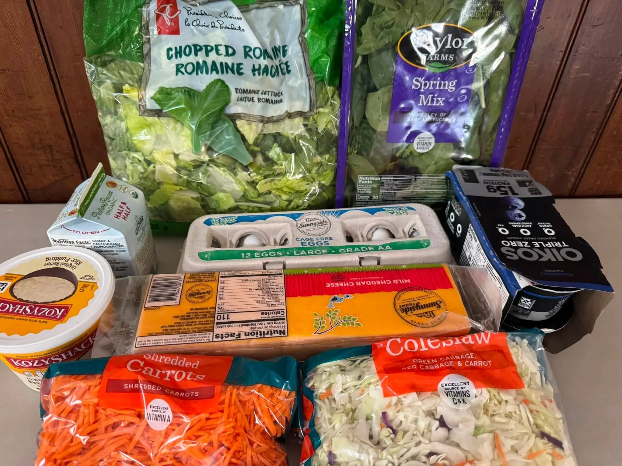 Assorted grocery items on a shelf including chopped romaine lettuce, spring mix, shredded carrots, chopped cabbage and carrots, eggs, yogurt, and almond milk.