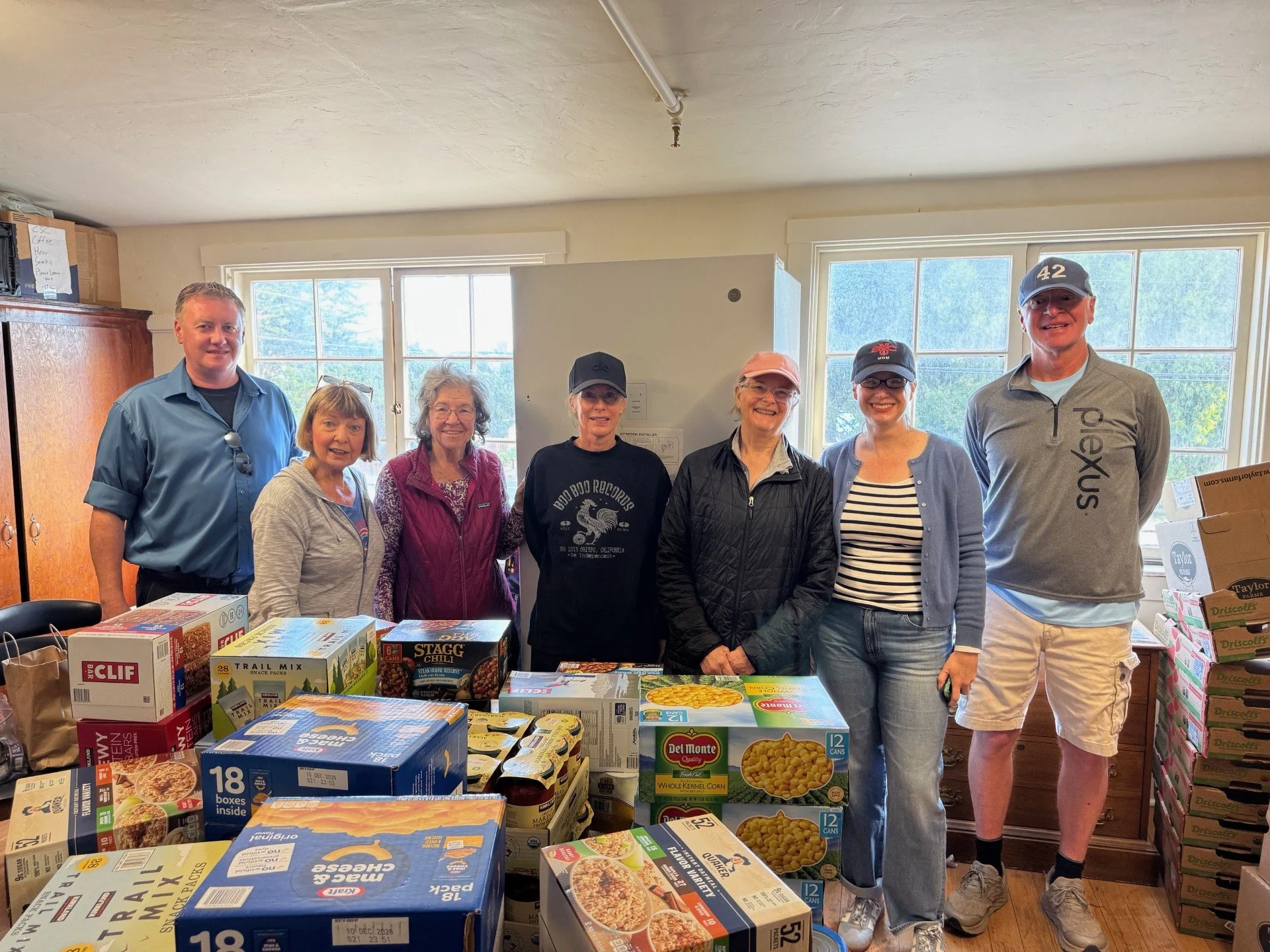 A group of seven people standing behind a table full of canned and boxed food items indoors, with large windows showing trees outside. The people are smiling and dressed casually.