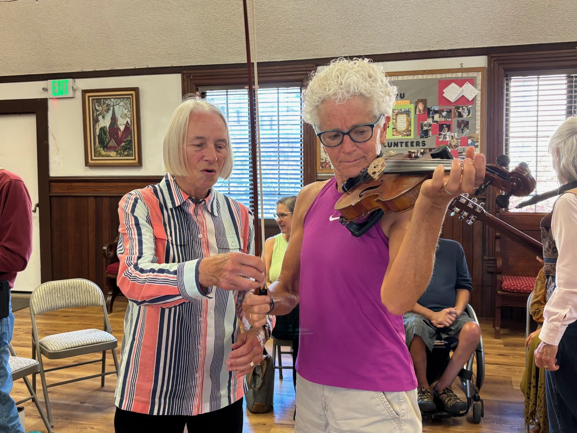 An elderly woman and a man with curly hair, glasses, wearing a pink tank top, playing a violin in a community room. Other people are seated in the background.