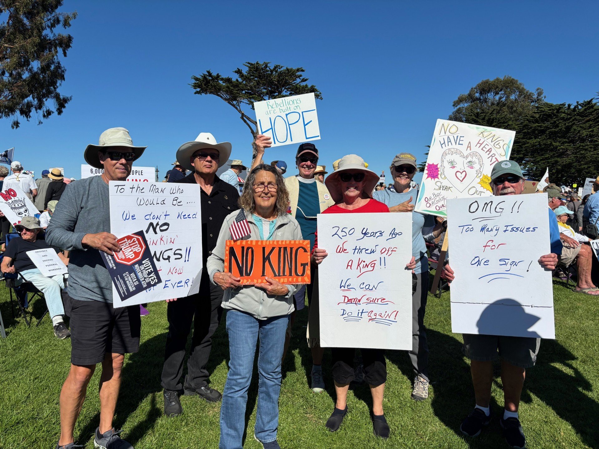 Group of protesters at a rally holding signs with various messages, including 'Rebellions are built on HOPE,' 'No KING,' and others addressing issues with monarchy, government, and social justice, outdoors on a sunny day.