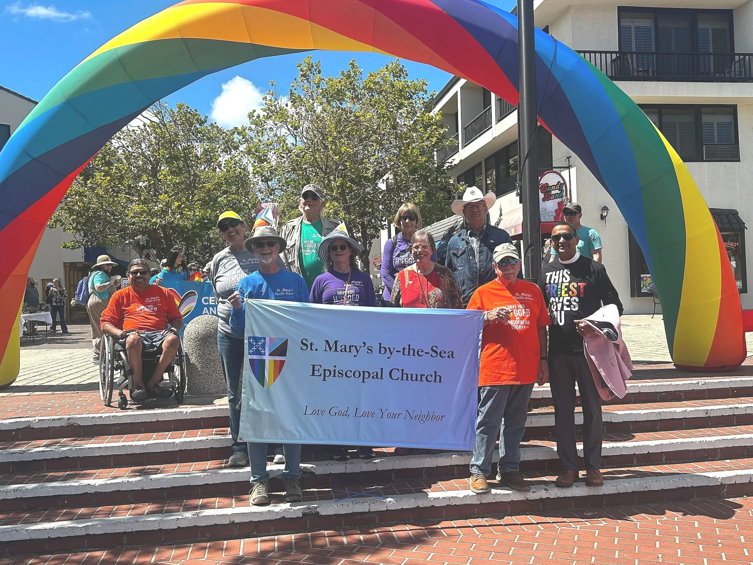 A group of people participating in a pride parade, holding a banner for St. Mary's by-the-Sea Episcopal Church. They are standing on brick stairs underneath a rainbow-colored arch. The crowd includes individuals of diverse ages and styles, some with hats and sunglasses, smiling for the camera. The background shows a building with balconies and trees.