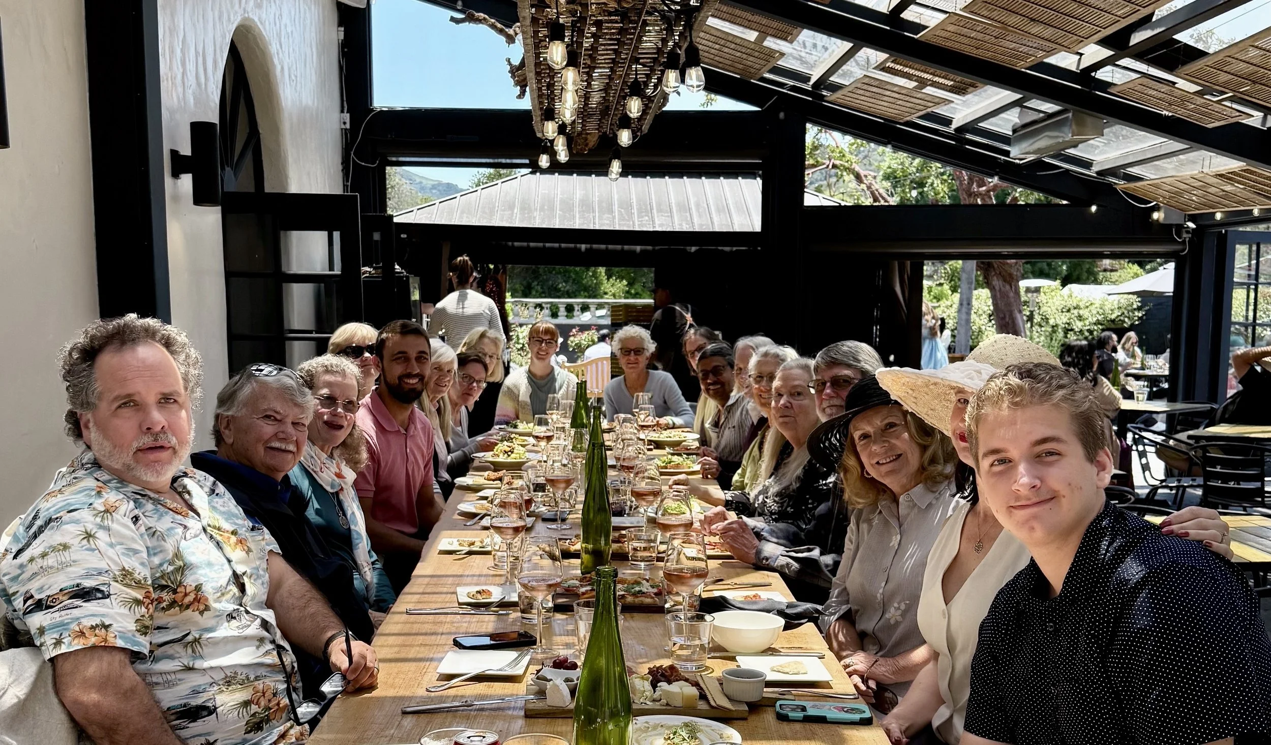 People gathered around a long dining table enjoying a meal at a restaurant with a glass roof and outdoor scenery in the background.